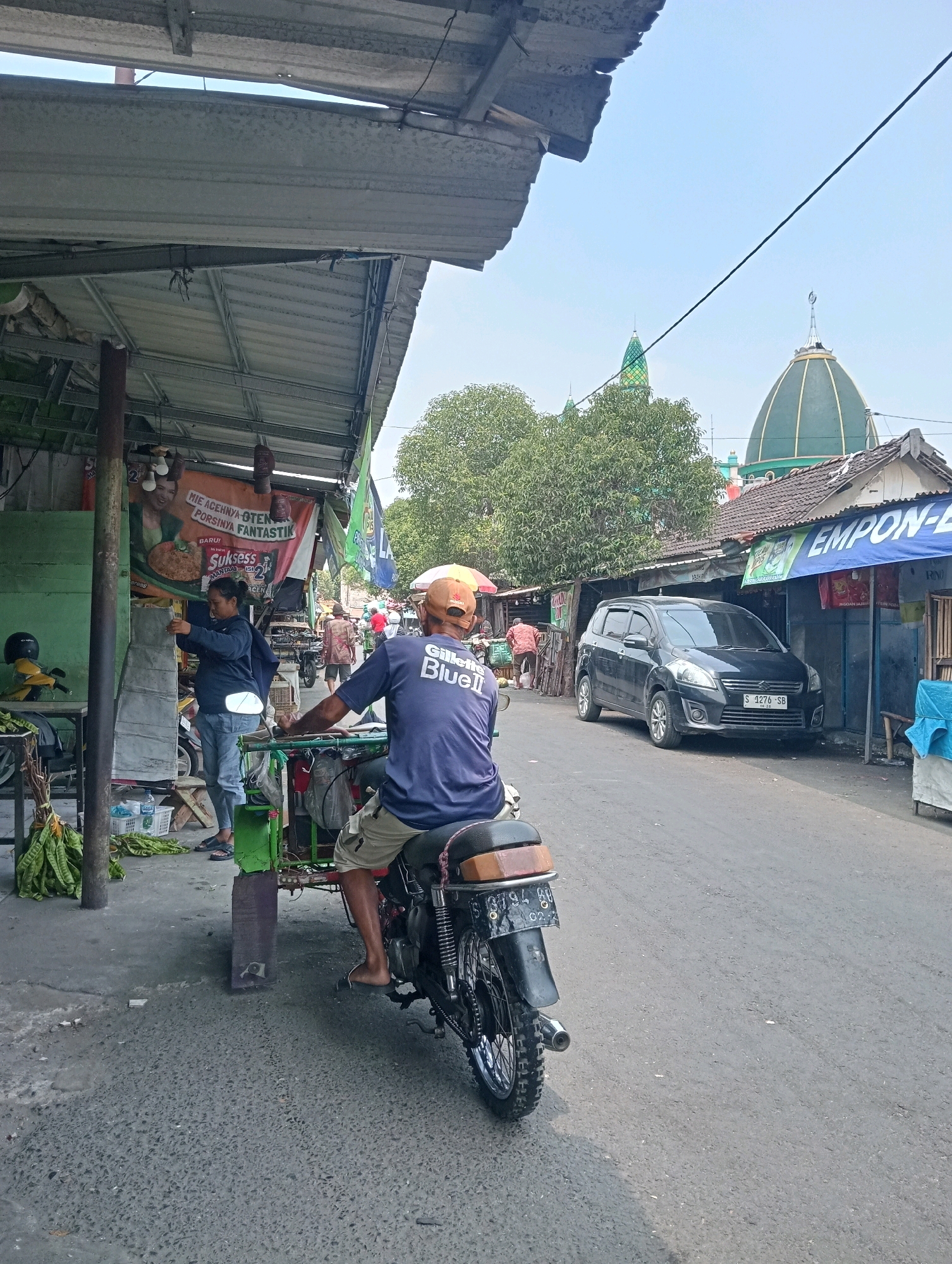 The image depicts a street scene in Jagalan, Indonesia. In the foreground, a person wearing a blue t-shirt with "Gillette Blue II" printed on it and a tan baseball cap rides a three-wheeled motorcycle. The motorcycle has a green and black frame and a black rear fender with a license plate that reads "S 3794 BG". The person is facing away from the camera. To the left, a woman in a blue jacket and jeans is standing and appears to be interacting with a display of what looks like food items. Behind her, a banner with Indonesian text is visible. Further down the street, a black car is parked. In the background, there are buildings with tiled roofs, trees, and two prominent green domes, suggesting the presence of a mosque. The sky is clear and blue. The environment appears to be a market or a commercial street with various stalls and vendors.