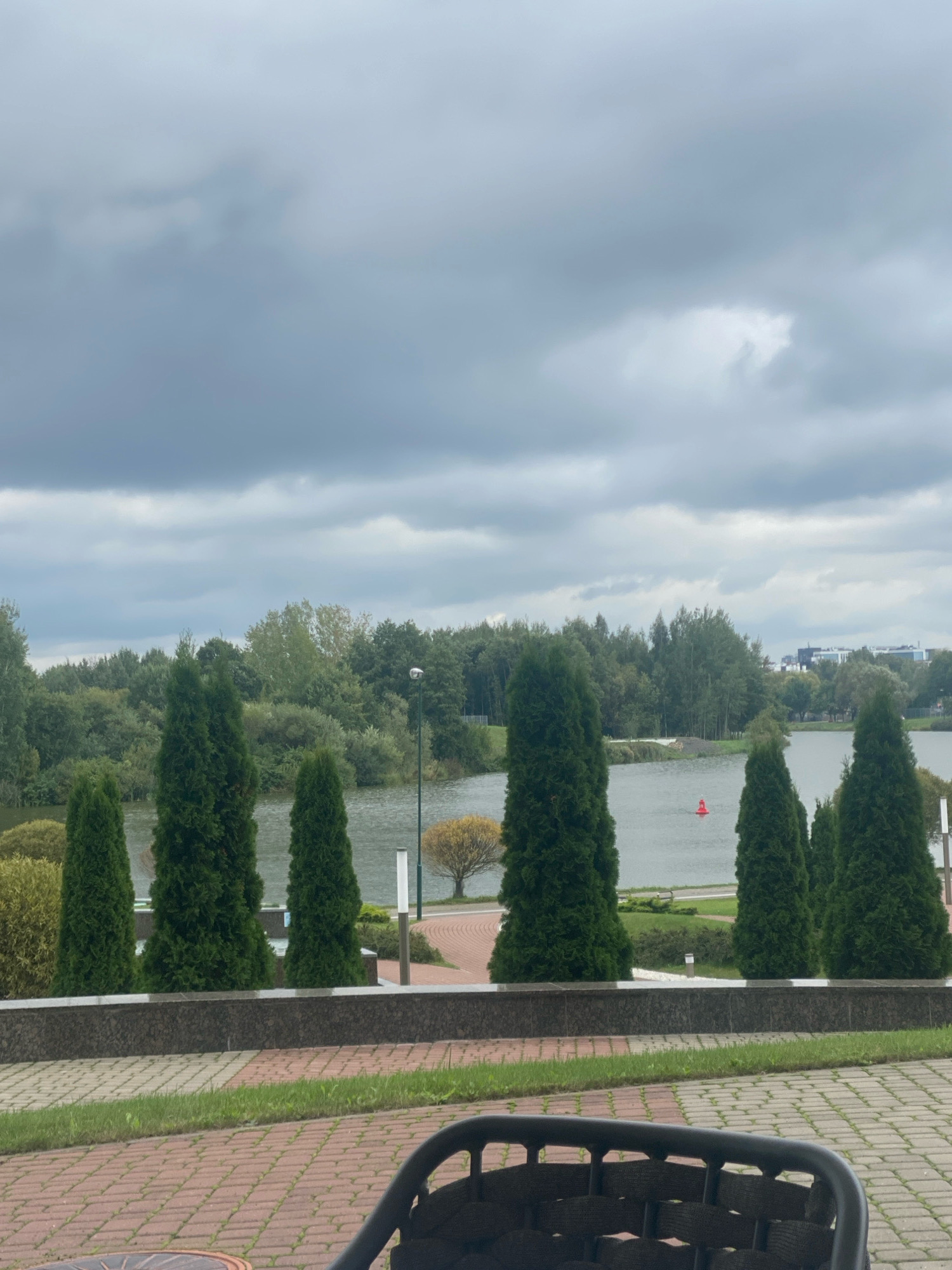 A cloudy day unfolds over a serene park setting in Minsk, Belarus, featuring a body of water and lush greenery. The foreground is occupied by a partially visible dark-colored outdoor chair, suggesting a seating area for visitors. Beyond the chair, a paved walkway with brick and stone patterns leads to a low, dark-colored granite wall.
A line of tall, conical evergreen trees stands in front of the water, creating a natural barrier. The water itself reflects the overcast sky, with a single red buoy visible in the distance. Across the water, a dense line of deciduous trees forms a verdant backdrop, with buildings faintly visible on the far right side of the frame, indicating the proximity of urban development. A lamppost stands near the water's edge, adding to the park's amenities. The overall atmosphere is calm and tranquil, typical of a park designed for relaxation and enjoyment.