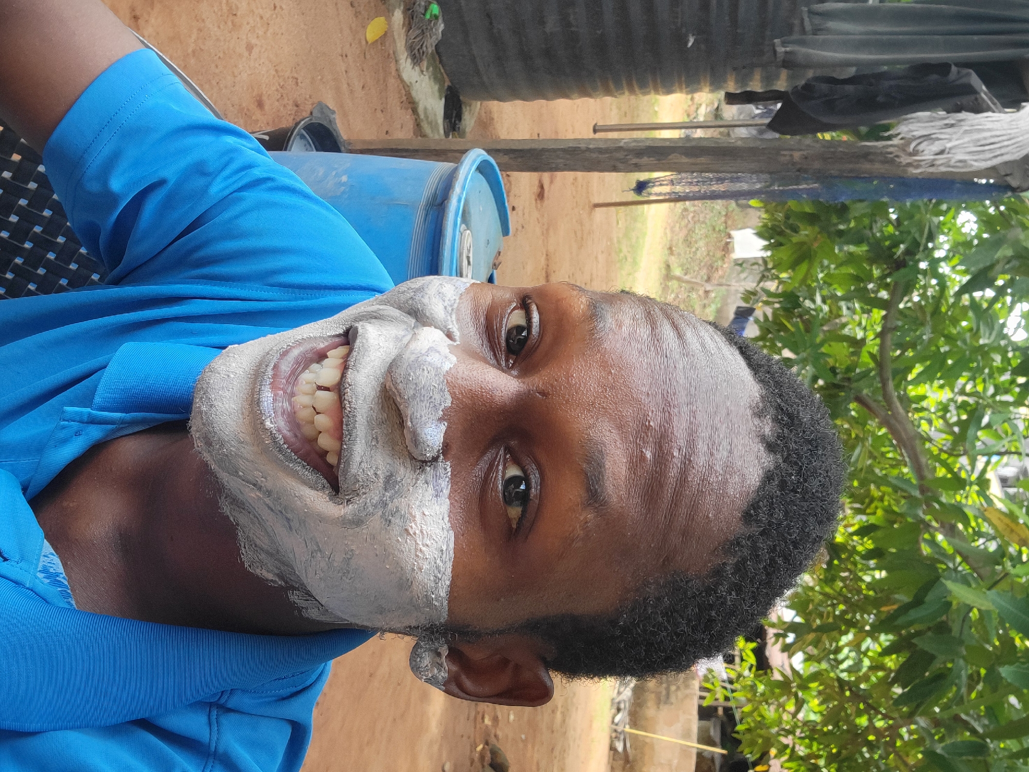 A man is centered in the frame. His face is partially covered with a thick, white, mask-like substance around his mouth and nose. He's smiling, revealing his upper teeth. He has dark skin and short, dark hair. He is wearing a blue shirt.
Behind him, there is a blue barrel, a wooden structure, some hanging clothes, and green foliage, suggesting an outdoor setting. The ground appears to be dirt or reddish soil.
Given the prompt, the location is Nkpor, Nigeria.