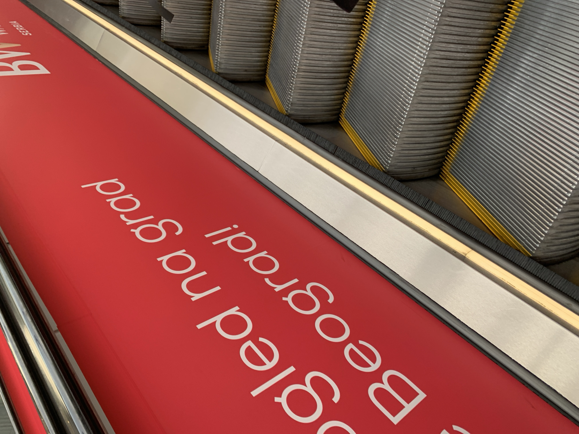 An upward-sloping view captures an escalator in what appears to be an indoor public space, likely an airport, shopping mall, or metro station in Belgrade, Serbia. The scene is dominated by a vibrant red advertising banner laid flat on the escalator's side panel, stretching from the lower left to the upper right.
No people are visible in the image. The main subjects are the mechanical components of the escalator: the steps, the handrail, and the advertising banner. The escalator steps are made of grey ridged metal, with bright yellow safety comb plates at their edges. A black brush strip runs along the base of the metal side panel. A polished chrome handrail is visible in the lower-left corner, and a segment of the metal trim with a narrow light strip runs along the top edge of the red banner.
The lighting suggests an indoor setting, likely from overhead artificial sources, as there are no shadows indicative of natural daylight. The time of day is not discernible.
The prominent white text on the red banner, when read upright, says "Dobrodošli na Beograd!" (Welcome to Belgrade!). Above this, towards the top left of the banner, the words "By" and "SERBIA" are partially visible, suggesting a tourism campaign or official welcome message related to the country and city.