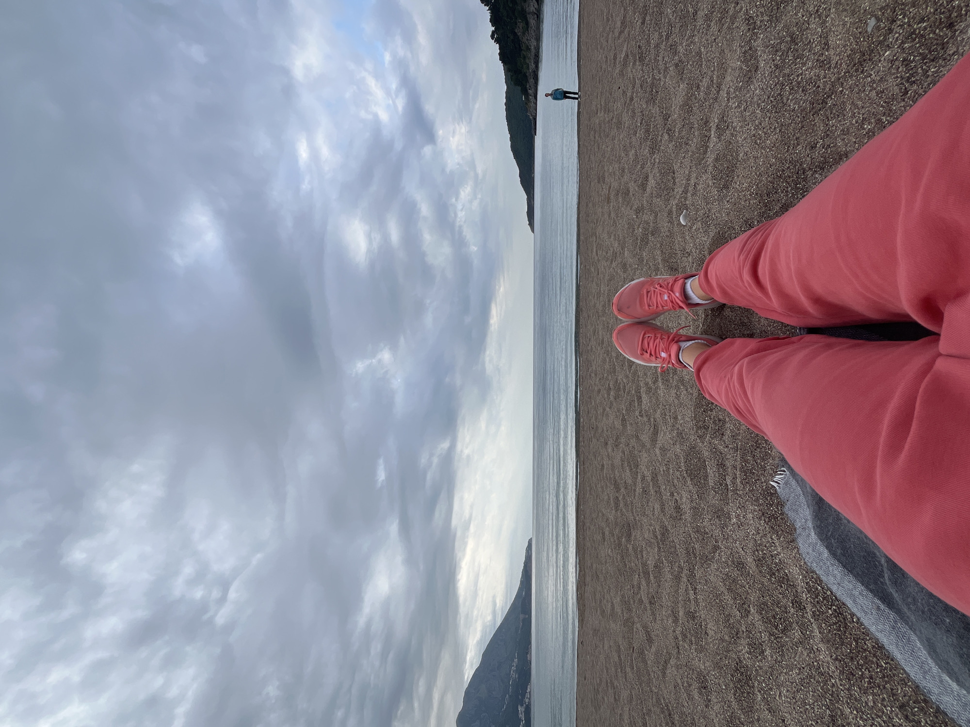 The photograph presents a beach scene, with the perspective being from the vantage point of someone seated on the sand. The foreground displays the lower portion of a person wearing red sweatpants and matching sneakers. The beach itself is comprised of dark sand.
The horizon line, where the sea meets the sky, bisects the image. The sea appears calm and reflects the overcast sky above. On the opposite side of the sea, along the distant shoreline, are the silhouette of mountainous terrains, possibly located in or near the city of Boreti, Montenegro. The sky is dominated by a blanket of grey clouds. In the background, a solitary figure stands on the beach.