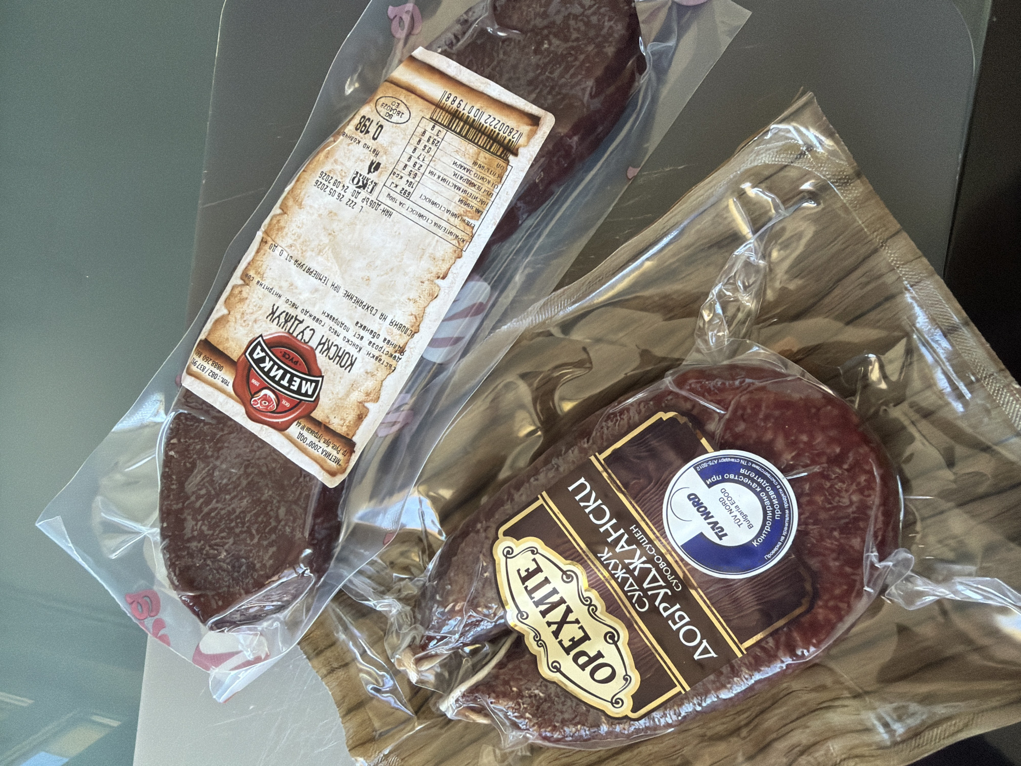 The image shows two vacuum-sealed packages of cured meat products on a dark, flat surface. The package in the foreground contains a dark red, irregular-shaped cured sausage labeled "ОРЕХИТЕ СУДЖУК ДОБРУДЖАНСКИ СУРОВО-СУШЕН". A round blue and white sticker with "TÜV NORD Bulgaria EOOD" is visible on this package. The package in the background contains a darker, more uniform slice of meat labeled "МЕТИКА КОНСКИ СУДЖУК". The labels are in Cyrillic script. The lighting suggests an indoor setting. There is no indication of the city of Dolno Ablanovo, Bulgaria in the image.