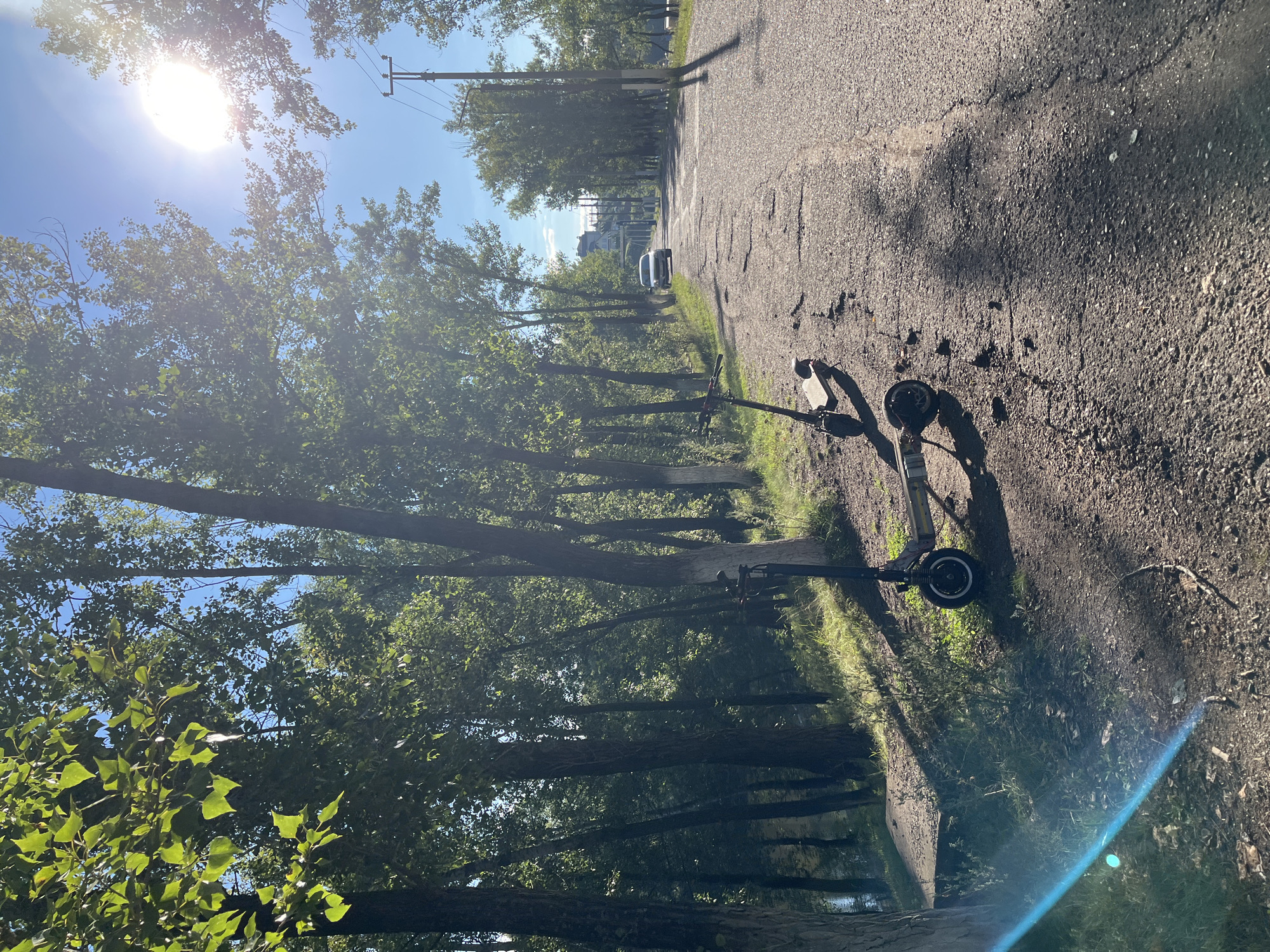 The image depicts an outdoor scene along a paved road bordered by trees and a body of water. Two electric scooters are parked on the side of the road, with one in the foreground and another slightly behind and to the right. The road appears cracked and weathered. Sunlight streams through the trees, creating dappled shadows on the ground. In the distance, a silver car is visible driving on the road. A utility pole with wires stands on the right side of the road. The environment suggests a park or a road near a natural area, possibly in Blagoveshchensk, Russia, given the context. There are no people visible in the image.