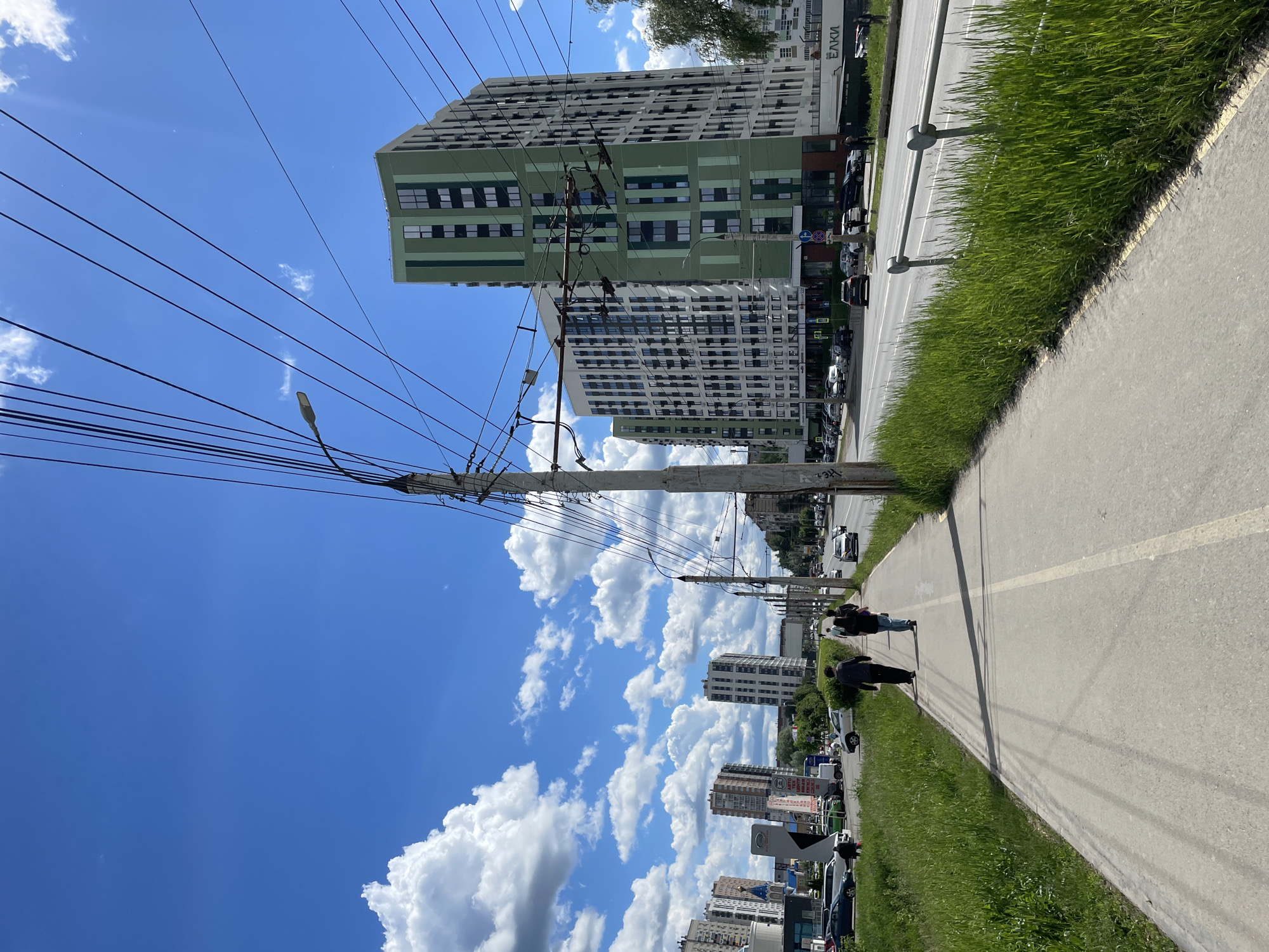 The image depicts an urban scene in Izhevsk, Russia, characterized by a wide pedestrian walkway alongside a road with vehicle traffic. Two individuals are walking on the walkway. One person is wearing a dark jacket, while the other is wearing a lighter shirt and darker pants. Parallel to the sidewalk is a strip of green vegetation separating the walkway from the street. A modern, high-rise residential building with a distinct green-and-white facade stands in the background, with "ELKI" visible on one side of the building. Overhead power lines and a utility pole are prominent in the foreground, intersecting the blue sky with cumulus clouds. Several vehicles, including cars and vans, are parked or moving along the street, indicating active urban activity. The weather appears clear and sunny. Other buildings of varying heights and architectural styles are visible further down the road, creating a cityscape backdrop.