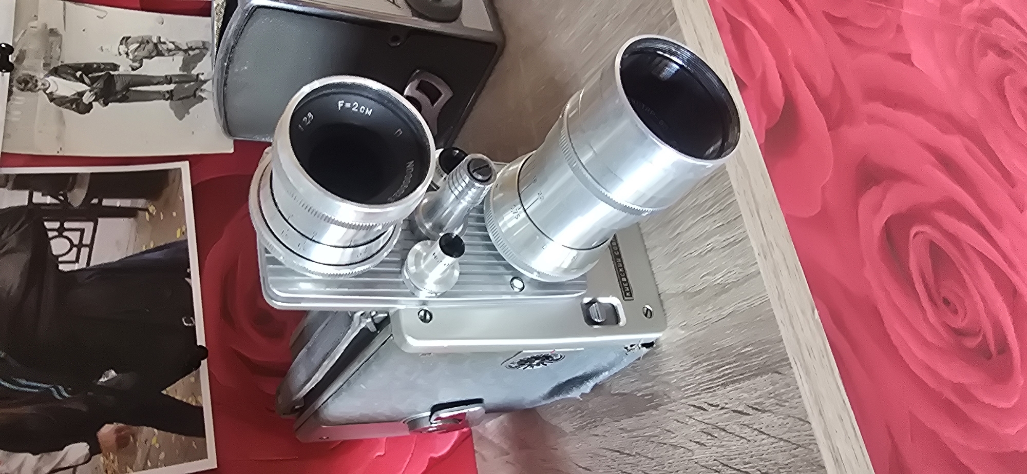 The image shows a vintage film camera on a wooden surface. The camera is silver and has a large lens. Behind the camera, there are two framed photographs. The photograph on the left shows a person in dark clothing. The photograph on the right shows a man and a child. The background is a red fabric with a rose pattern. This scene is located in the city of Gostagayevskaya, Russia.