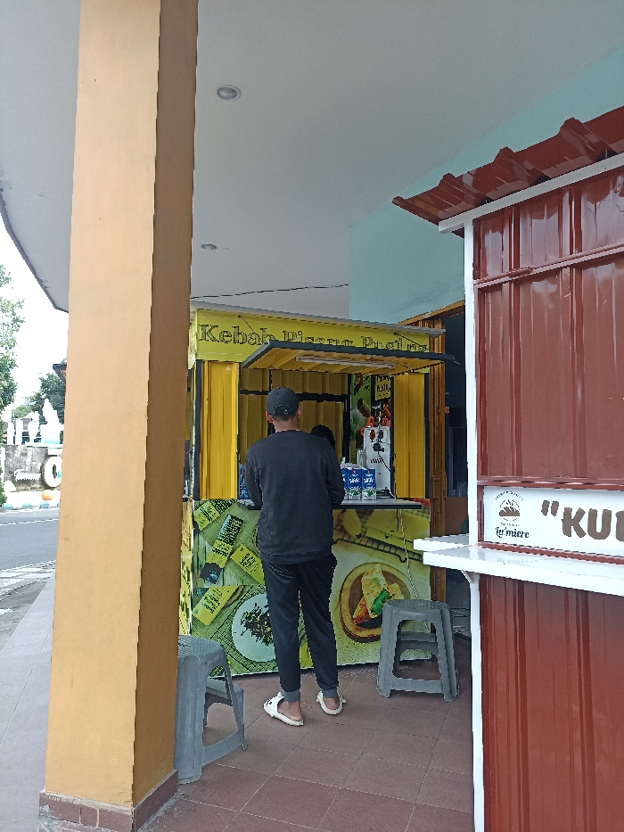 The image shows a daytime scene featuring a man standing at a food stall. The man, viewed from behind, wears a dark long-sleeved shirt, dark pants, a baseball cap, and white sandals. He is positioned facing a bright yellow food kiosk, suggesting he is ordering or waiting for food.
The yellow kiosk is constructed from corrugated metal and has an open serving window. Text on its top reads "Kebab Pisang Pra...", and its lower part is decorated with graphics of various food items, including what appears to be a wrapped dish. On the counter of this kiosk, several white rectangular containers, possibly milk cartons (one visible brand resembles "ULTRA MILK"), and some machinery are present. Two grey plastic stools are placed in front of the stall.
To the right, a second food stall, made of brown corrugated metal, is partially visible. It features a white serving counter, with the text "Le miere" and a partial word "KUL" displayed on its front.
The scene takes place under a covered area, indicated by a light-colored ceiling and a large ochre-colored pillar on the left. In the background, a paved road, some white structures, and greenery are discernible, suggesting a street or public space. The lighting indicates it is daytime. The overall impression is that of a casual outdoor food vendor setup, common in locations like Tulungagung, Indonesia.