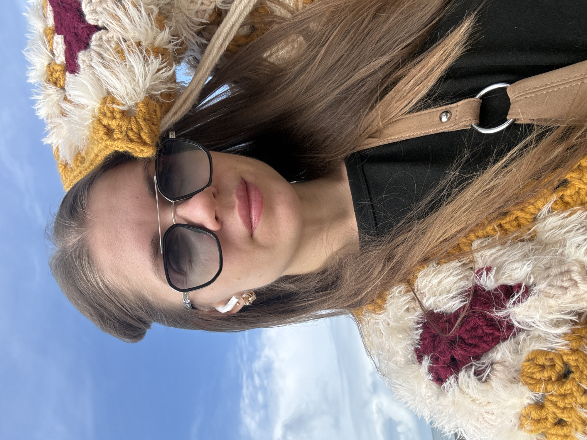 A woman is photographed outdoors against a sky with white clouds and blue sky. She is wearing sunglasses, a multicolored knitted or crocheted hat and scarf, and a black shirt. A tan strap or handle is visible over her right shoulder. She has long brown hair and is wearing a small earring in her left ear. It is not possible to tell that the photograph was taken in Batumi, Georgia.
