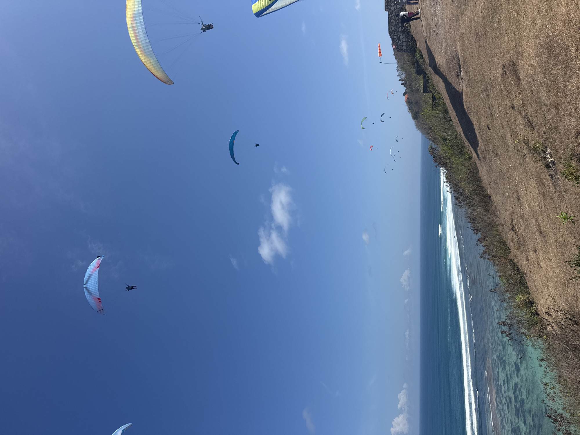 In an open sky above a coastal landscape, several paragliders are visible. The sky is a clear, vibrant blue, with a few scattered white clouds. The paragliders, with colorful canopies, are suspended in the air, appearing to be in mid-flight. The landscape below consists of a grassy hill sloping down towards a turquoise ocean. Waves are breaking along the shore, creating a white foam line. In the distance, there are some buildings and structures visible on the coastline, including what appears to be a stone wall or barrier. A few people are standing near the structures, observing the scene. The overall impression is one of a recreational activity taking place in a scenic, coastal environment.
