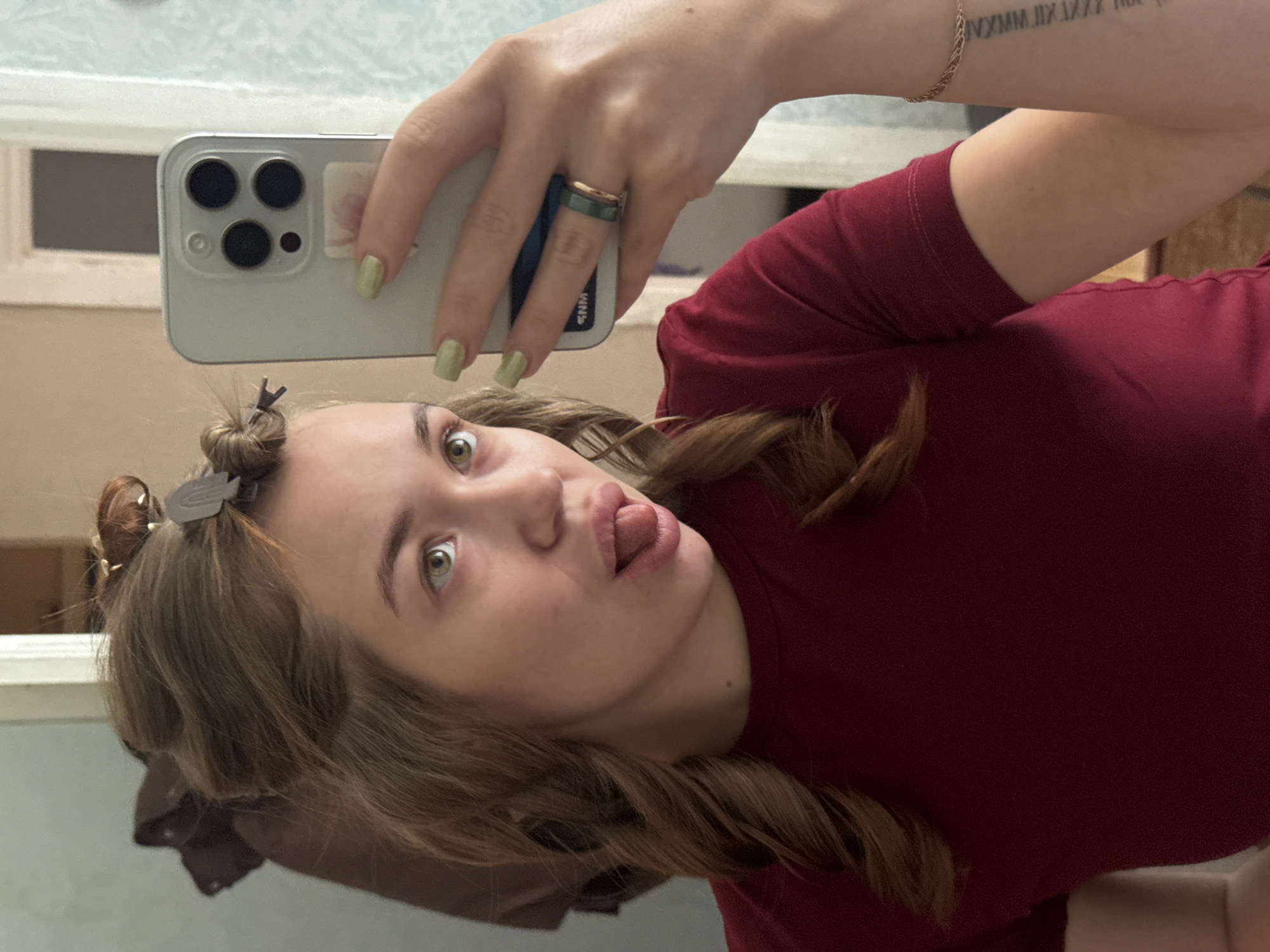 In a close-up selfie taken in what appears to be a bathroom, a young Caucasian woman with light brown hair styled in rollers is looking at the camera with her tongue sticking out. She is wearing a dark red t-shirt and her left arm, adorned with a bracelet and a Roman numeral tattoo, is extended, holding a smartphone. A ring with a green band and wooden inlay is visible on her left ring finger. The background suggests an indoor setting, possibly a home, with a mirror reflecting part of her face and hair. The image was captured in Chaikovsky, Russia.
