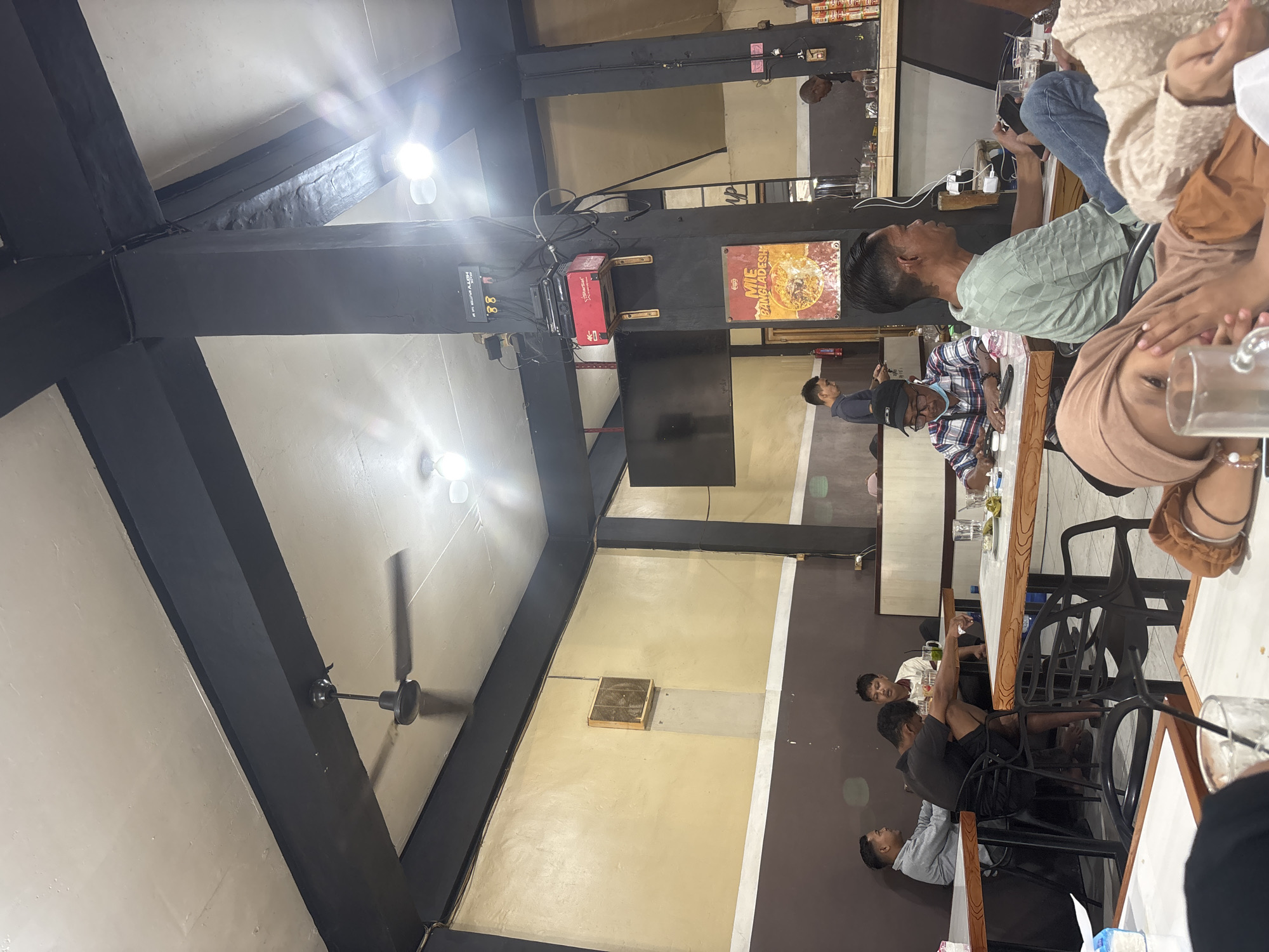 The image shows the interior of what appears to be a restaurant or dining establishment. The location is Keuniree, Indonesia, based on the "MIE BANGALADESH" poster on the wall.
The interior features a high ceiling with black beams and white panels. Light fixtures and a ceiling fan are visible on the ceiling.
Several individuals are seated at tables, engaging in conversation and likely dining. At least one person is wearing a hijab. Other patrons are also present in the background. There are glasses and other dining items on the table in the foreground. The overall atmosphere seems relaxed and casual.