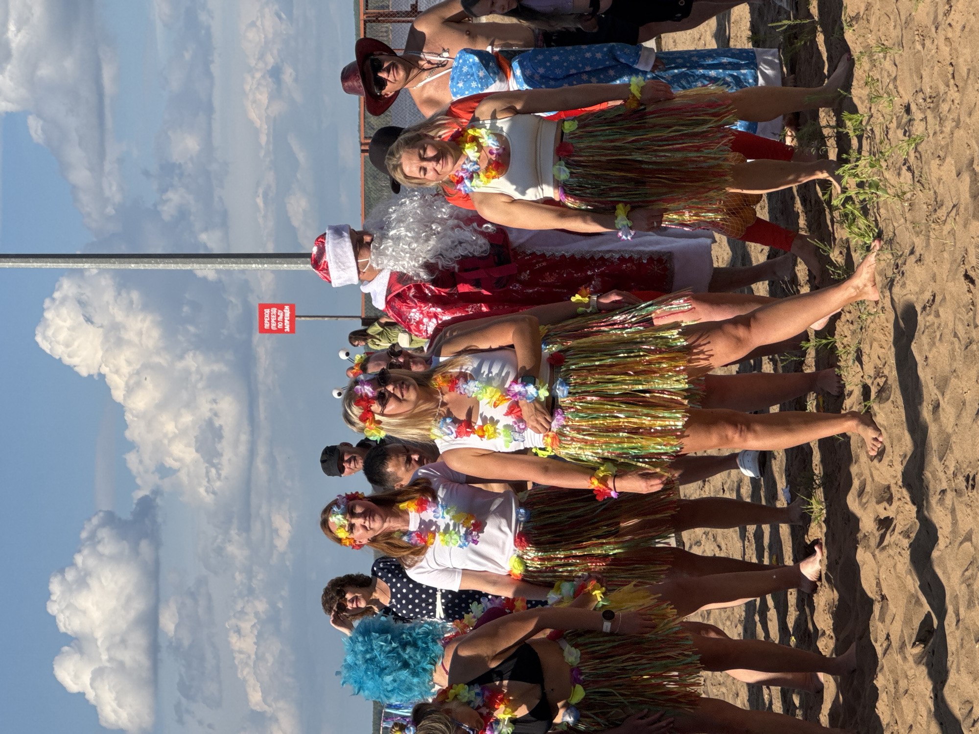 In the city of Chaikovsky, Russia, on a sandy beach under a partly cloudy sky, a group of people are gathered. The foreground is dominated by the legs and lower torsos of several individuals. Many are wearing colorful, shiny grass skirts, and some have leis around their necks or as wristbands. One person in the front left has bright blue, spiky hair. Behind them, a woman in a black and white polka dot dress stands out, shielding her eyes from the sun. Further back, a person dressed as Santa Claus, complete with a red suit, white beard, and hat, is visible. To Santa's right, a man wearing a cowboy hat and sunglasses is present. The background shows a metal fence and some distant greenery. A red sign with white lettering is attached to a pole. The overall impression is of a festive gathering, possibly a themed event or celebration.