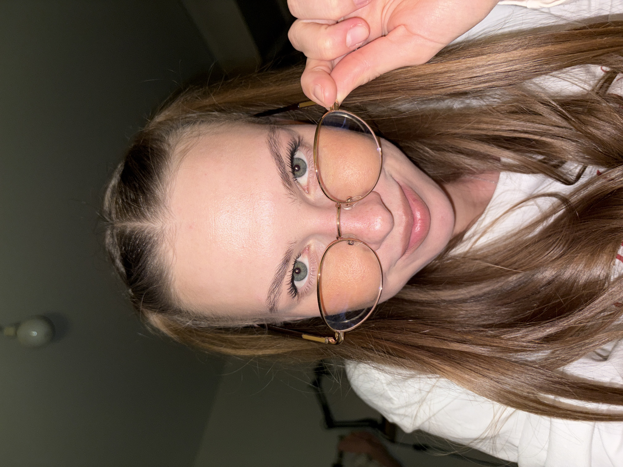 A young woman with long, brown hair styled in pigtails is looking at the camera while holding her glasses. She is wearing a white t-shirt. The background is dark and appears to be an indoor setting. There is a lightbulb visible on the ceiling in the upper left corner of the image. The scene is a close-up shot of the woman's face and upper body. There is no identifiable information in the image to suggest a location in the city of Perm, Russia.