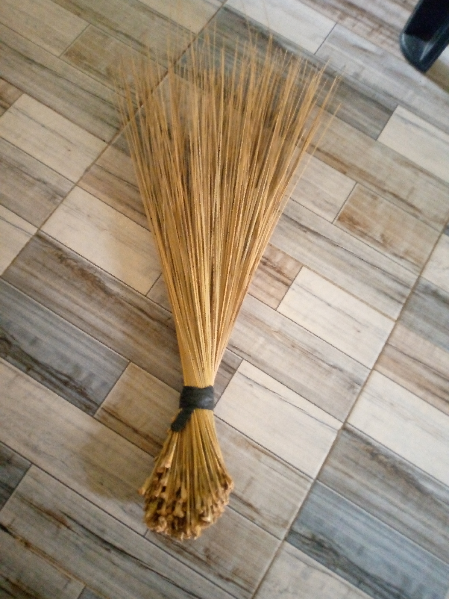 A broom made of dried grass is lying on a floor of grey and beige wooden planks, arranged diagonally. The broom's handle is tied with a black band. The broom is positioned at an angle, with the bristles fanned out and pointing towards the top right of the image. The floor appears to be indoors, likely in a residential or commercial setting. The image does not contain any people or indicate a specific location within Jalingo, Nigeria. The lighting suggests the image was taken during the day.