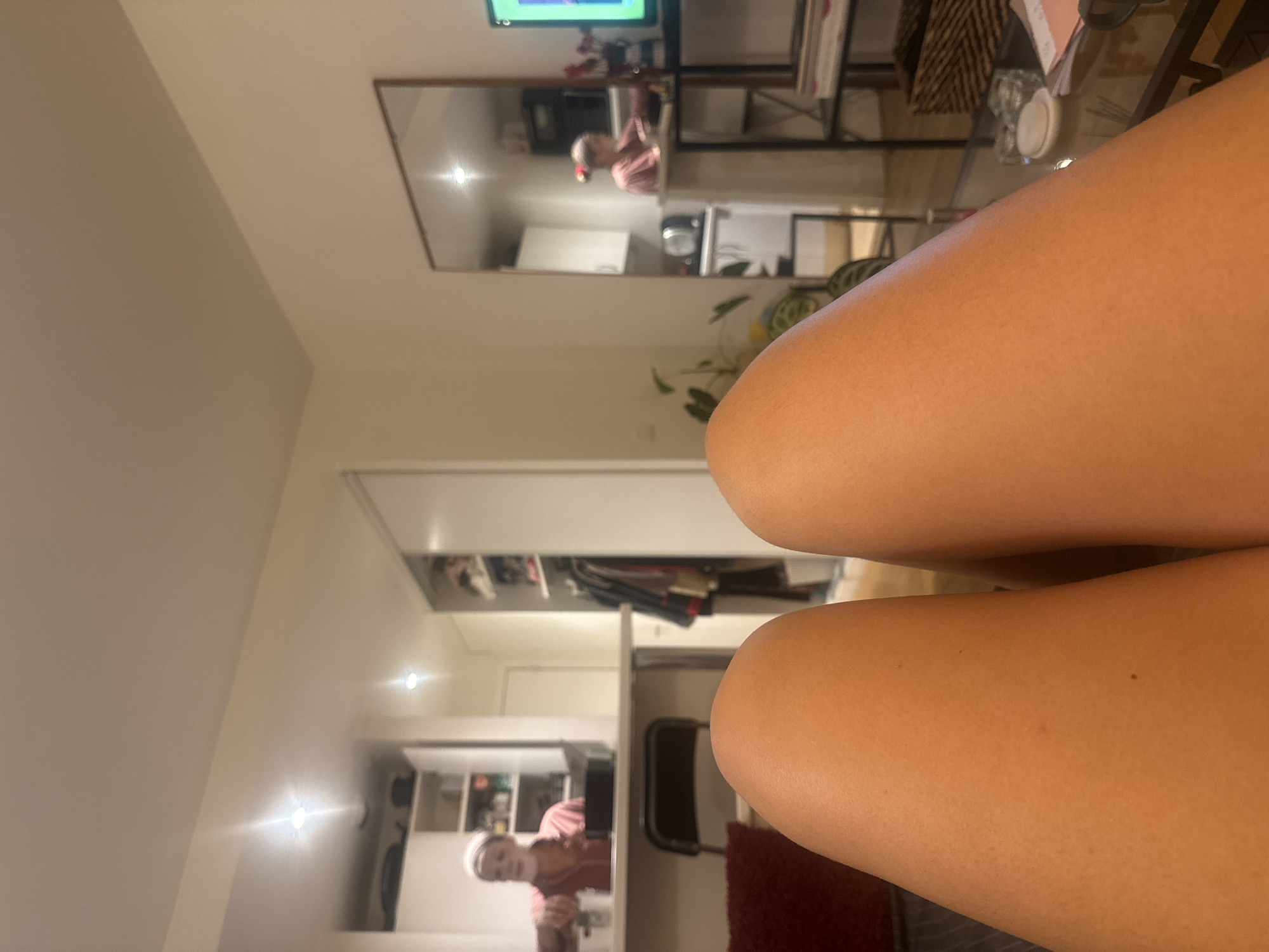 The image shows a pair of legs in the foreground, obscuring a distorted view of what appears to be a room. The room is visible through a mirror reflection and features a kitchen area with white cabinets and a person standing in front of a counter. There are also elements of a living area, including a television and a bookshelf. The lighting is warm, suggesting evening or indoor illumination.
The image does not contain enough details to confidently determine if it is in Paris, France.