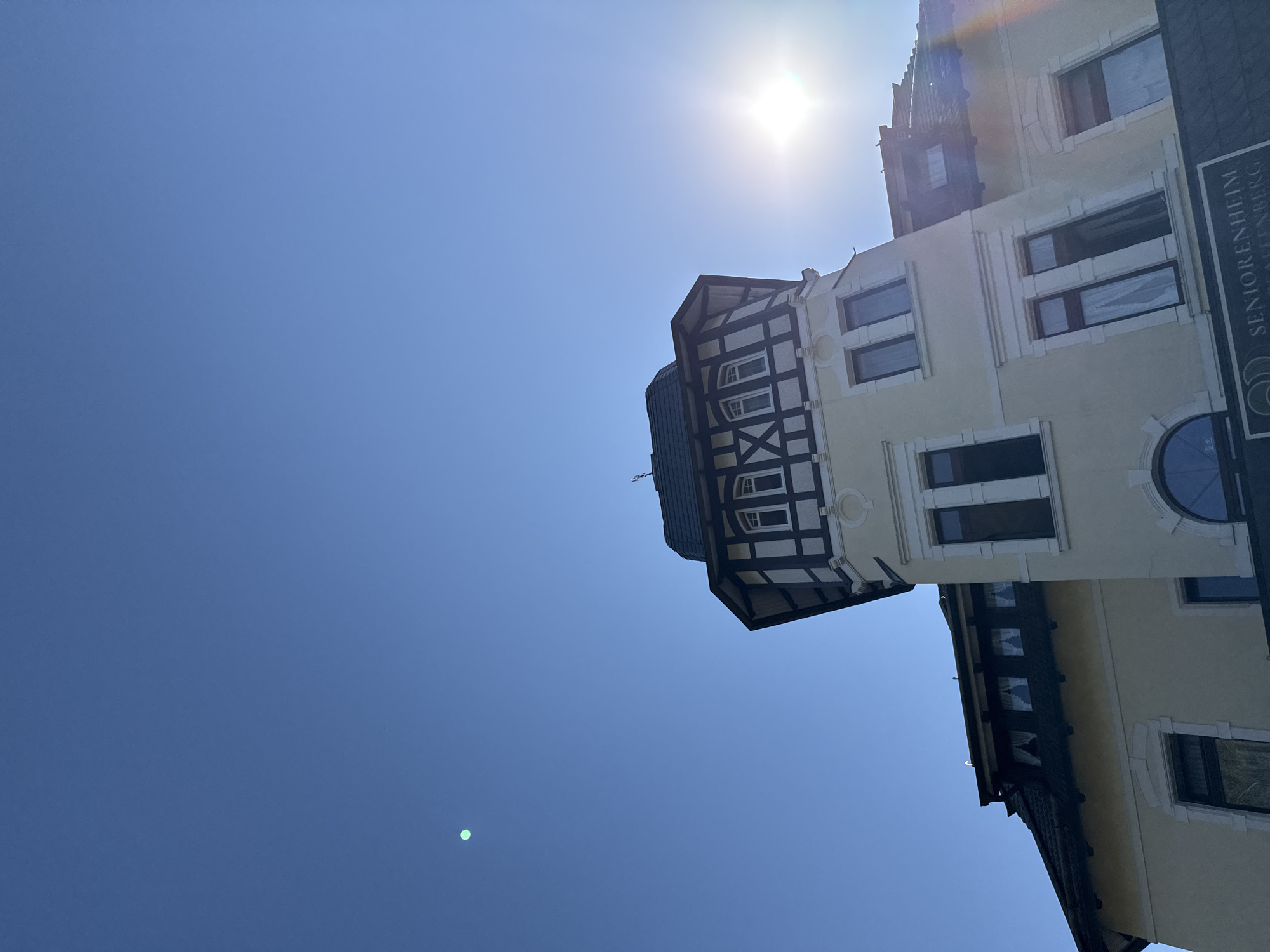 This is a low-angle, full-shot photograph of a building in the city of Bad Sachsa, Germany. The building is a multi-story structure with a prominent tower that features a half-timbered upper section. The tower has a dark, sloped roof with dormer windows. Below the tower, the building is painted a light yellow with white trim around the windows and decorative architectural elements. There are several windows visible on the facade, some with curtains. A sign at the bottom of the frame reads "SENIORENHEIM" and "GRAFENBERG". The sun is shining brightly, creating a lens flare in the upper right portion of the image. The sky is a clear, deep blue, with a small, faint green circular anomaly visible in the upper left quadrant, possibly a reflection or lens artifact. The overall scene depicts a well-maintained building against a bright, sunny sky.