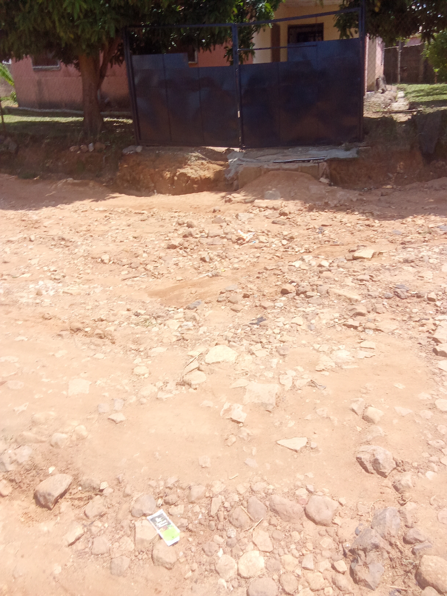 The image depicts a dusty, unpaved road in front of a dark metal gate, likely an entrance to a property. The ground is covered in a mixture of sand, small stones, and larger rocks. To the left of the gate, a tree with green foliage grows, and behind the gate, a building with a light-colored facade and a red-tiled roof is partially visible. The environment appears to be dry and somewhat neglected. A small, colorful object, possibly a piece of litter or packaging, is visible on the ground in the lower right portion of the image. There are no people or animals visible in the scene. The overall impression is of a rural or semi-urban area in an arid or seasonally dry climate, which could be consistent with parts of Jos, Nigeria.