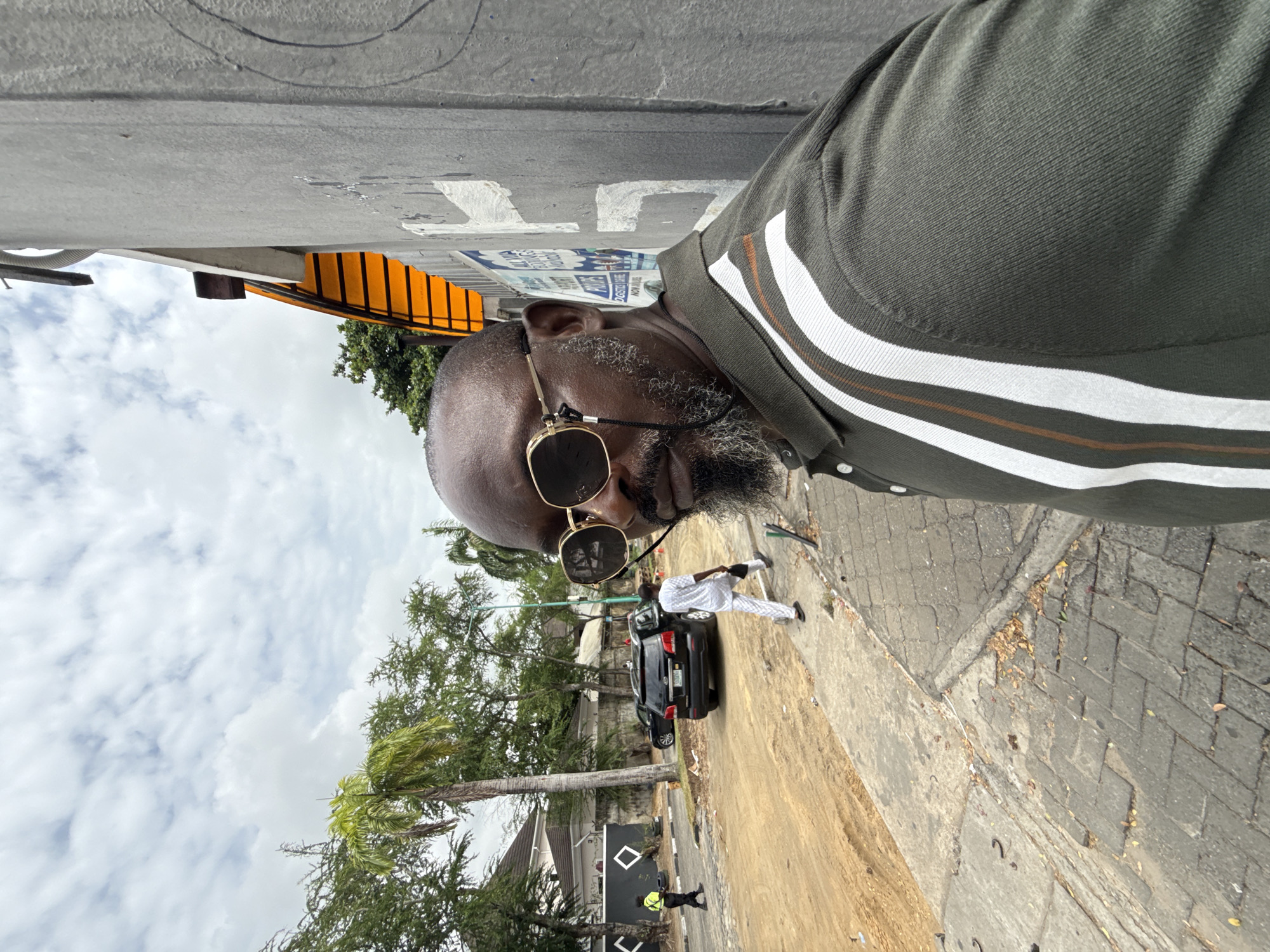The image is a first-person perspective shot taken in Lagos, Nigeria. A man wearing sunglasses and a polo shirt with white and brown stripes stands on a street with a sidewalk. A black car and a man in white clothes are in the background. Also visible is a yellow traffic barrier. The location appears to be a residential area with trees and a cloudy sky overhead. A person in a yellow vest is visible near a wall marked with white diamond shapes. The scene includes elements of both street activity and an urban environment.