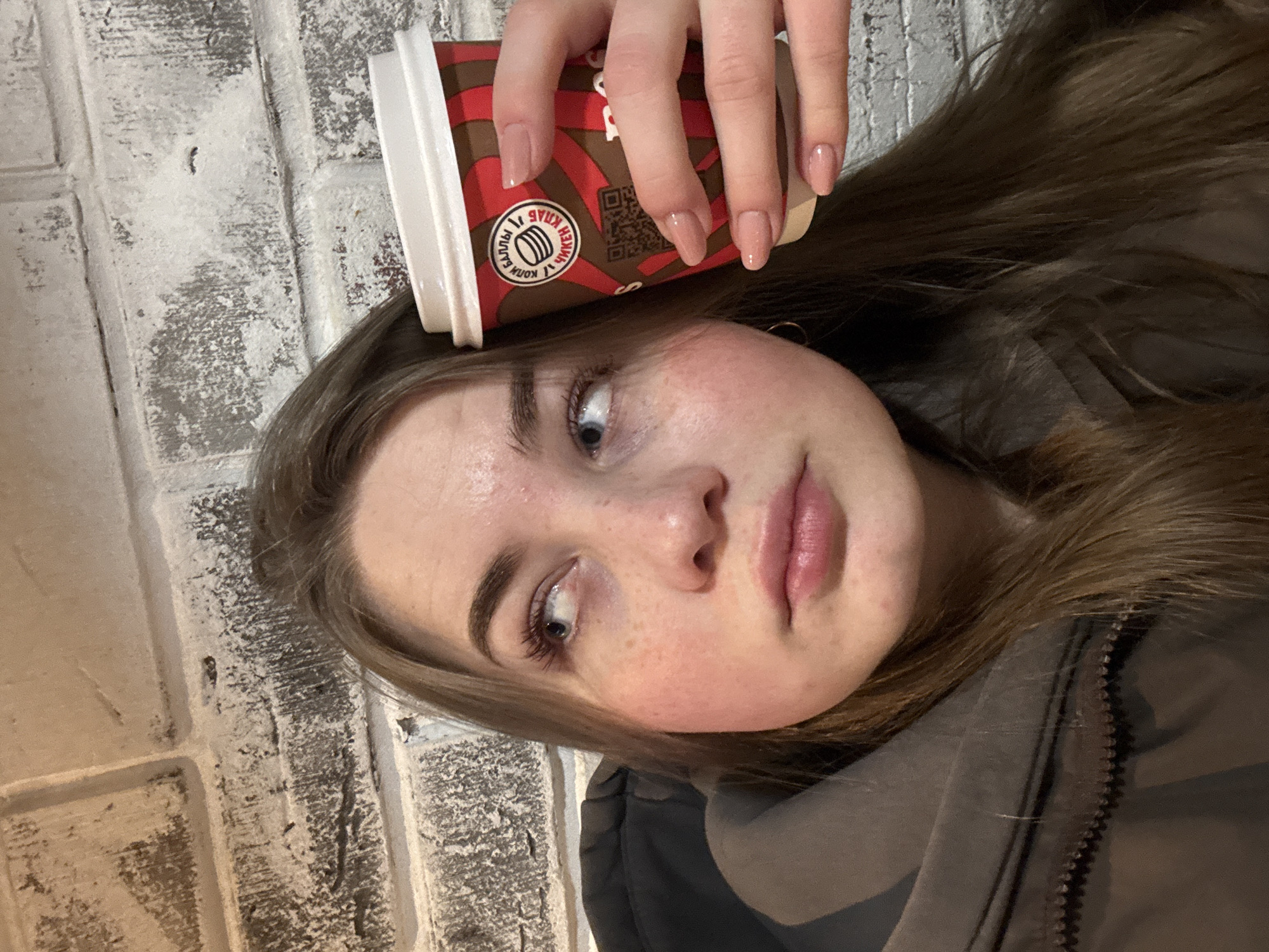 This vertically oriented image presents a close-up portrait of a young individual, likely female, captured from the chest up. She has light brown hair, fair skin with visible freckles on her cheeks and nose, and light-colored eyes. Her long eyelashes and well-defined eyebrows are notable. Her expression is neutral, with her gaze directed slightly upwards and to the side. She is wearing a dark, possibly grey or brown, zippered jacket.
Her hand is visible at the top of the frame, holding a red and brown disposable coffee cup, which is positioned casually on her head. Her fingernails are manicured with a light, neutral polish. The coffee cup features a distinctive design with patterns in red and brown. A circular logo on the cup displays Cyrillic text: "КУБОК СЧАСТЬЯ" (Cup of Happiness) and "ЧАЙ ИЛИ КОФЕ" (Tea or Coffee), along with a QR code.
The background is a white or light-colored brick or textured wall, suggesting an indoor or sheltered outdoor environment. The lighting is ambient, without strong highlights. The overall impression is candid and informal, depicting a moment in Chaikovsky, Russia.