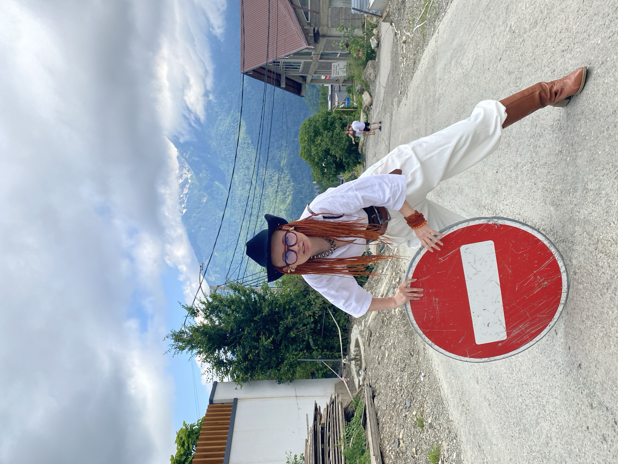 A woman with bright orange braided hair, wearing a black cowboy hat, glasses, a white unbuttoned shirt, white pants, and brown boots, poses with a red "no entry" sign in the city of Krasnaya Polyana, Russia. She is on a dirt road with scattered gravel. In the background, there are buildings with brown roofs, a white wall, stacks of lumber, trees, and a mountainous landscape under a cloudy sky. A man and a child are visible in the distance, further up the road.