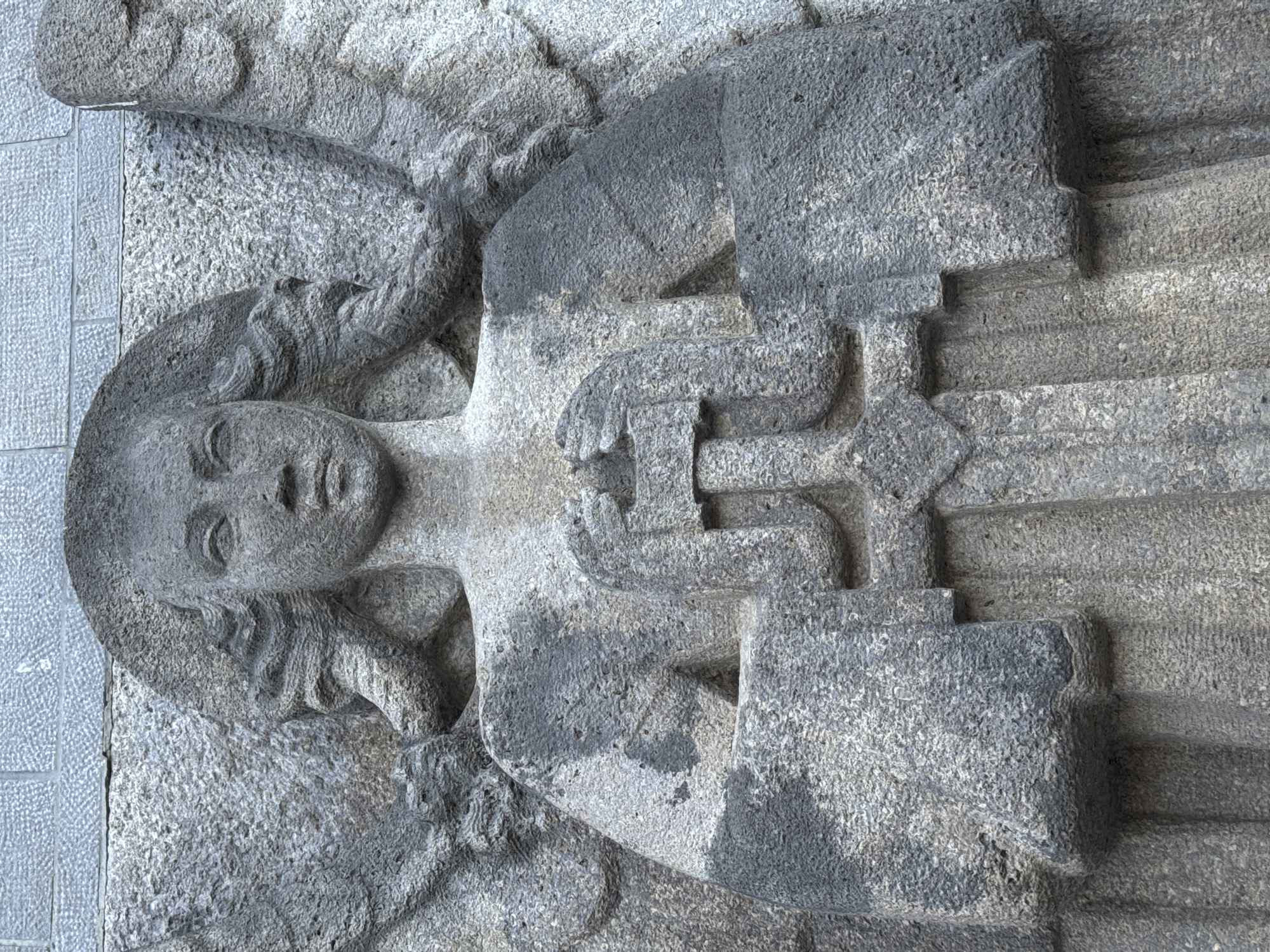 This is a close-up, eye-level, vertical, full-body, medium shot of a stone relief sculpture of an angel. The sculpture is carved from gray stone and depicts the angel from the chest up, with visible wings on either side. The angel has a serene facial expression with closed eyes and flowing hair. Its hands are clasped in front of its chest, holding a cross. The angel is wearing a long, draped robe. The background is a textured stone wall, possibly part of a building. The sculpture is located on the facade of the Church of Our Lady of the Angels (Onze-Lieve-Vrouwekerk) in Strombeek-Bever, Belgium.