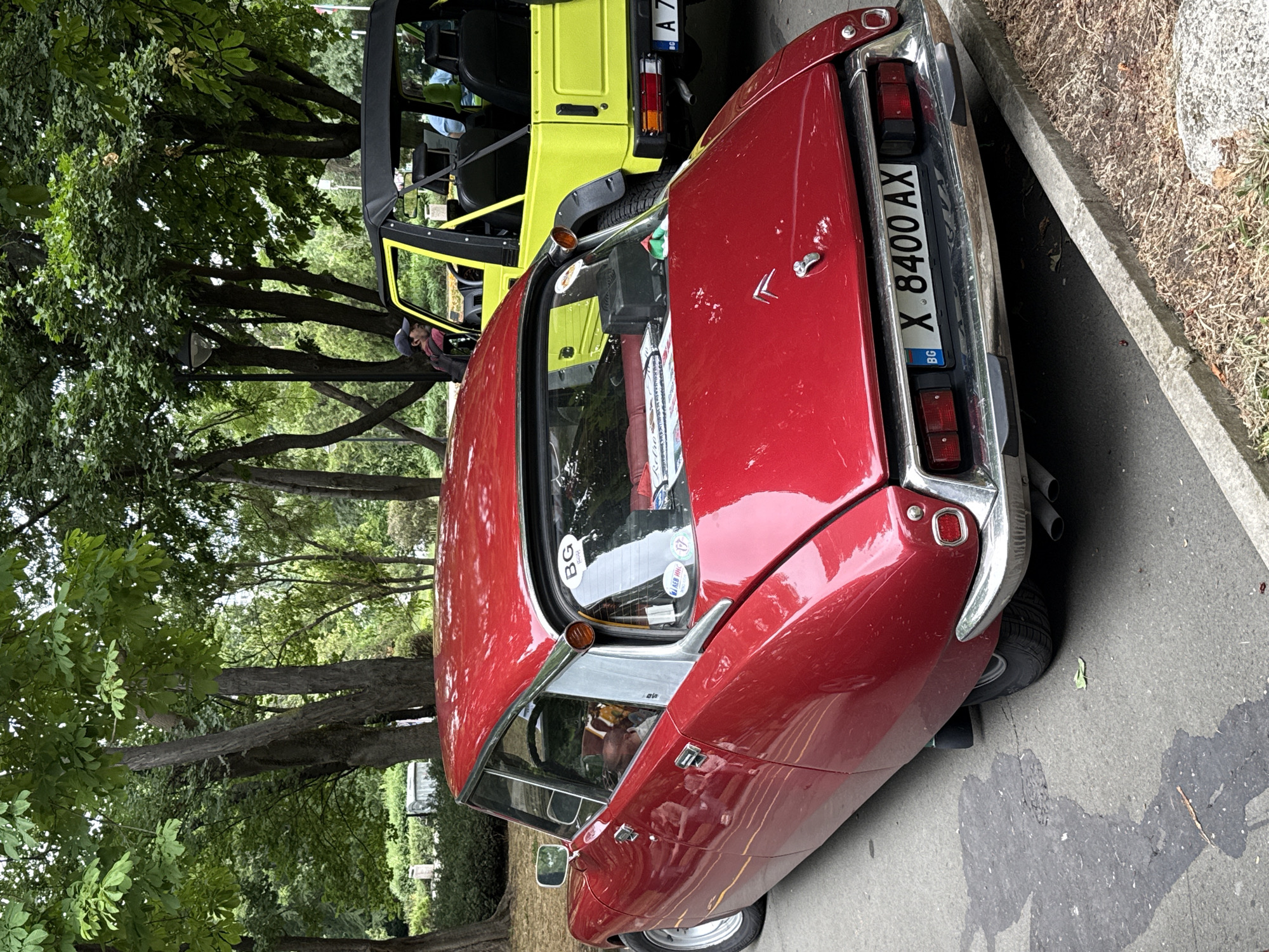 A red, vintage Citroen DS car is parked on a paved surface next to a curb and dry grass, with a bright yellow Suzuki Jimny parked behind it. Both cars are viewed from the rear. The Citroen has a license plate reading "BG X 8400 AX". The Suzuki has a license plate reading "A 7...". Trees with green leaves overhang the scene, indicating it's likely a park or a tree-lined street. The overall lighting suggests it's daytime. The scene appears to be in the city of Burgas, Bulgaria, potentially at a car show or exhibition given the presence of multiple vintage and distinct vehicles. There are no people clearly visible in the immediate vicinity of the cars, though a person can be seen in the driver's seat of the Suzuki, and another person is partially visible inside the Citroen.