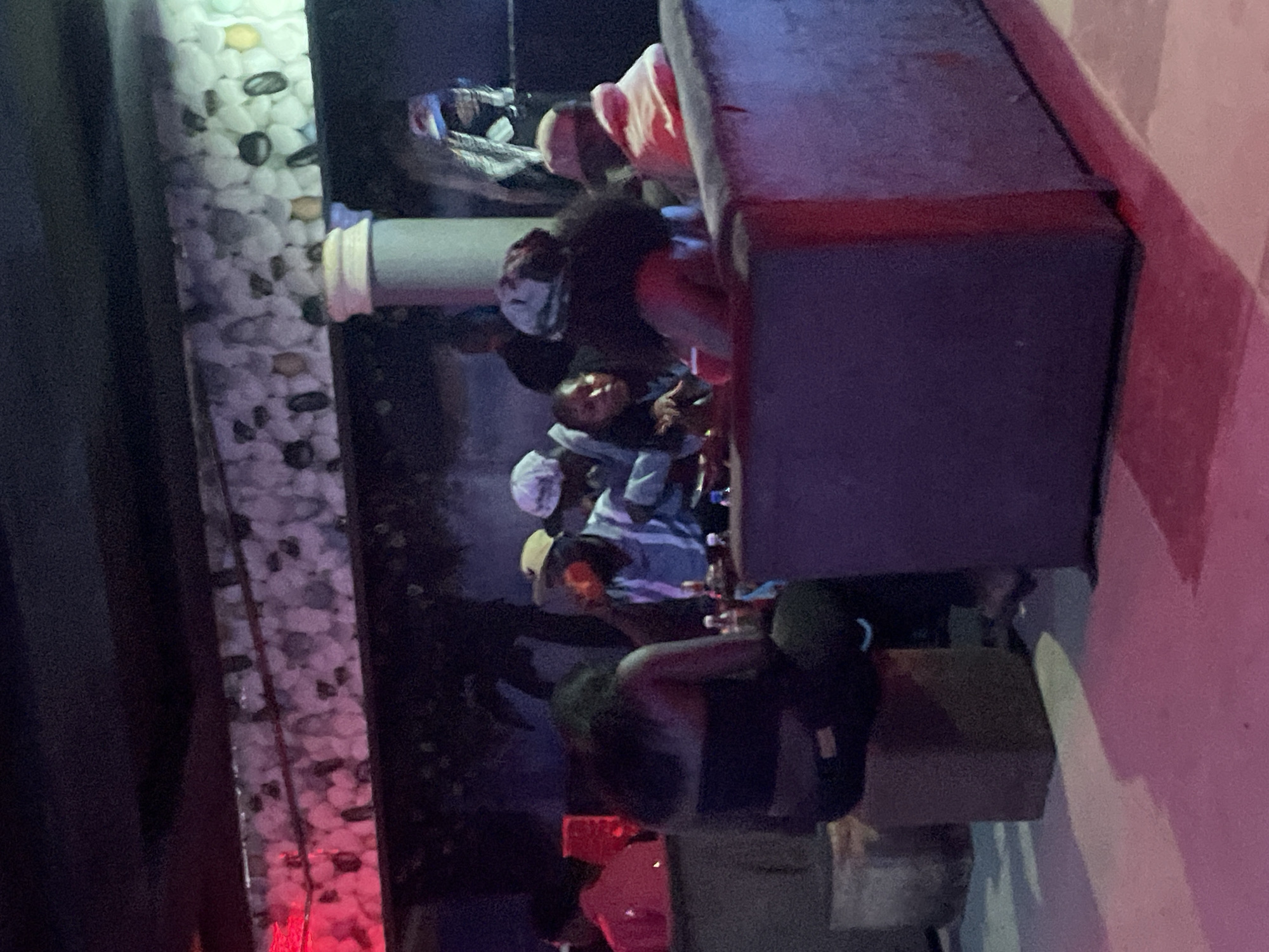In Durumi, Nigeria, a dimly lit establishment, possibly a lounge or club, hosts a gathering of individuals seated around tables. The lighting is subdued, with pink and purple hues illuminating the space. In the foreground, a dark-colored couch dominates the right side of the frame. To the left, a woman with dark curly hair is seated, facing away from the camera, wearing a black strapless top and black pants. A square stool sits in front of her.
Further into the scene, a group of people are engaged in conversation around a table laden with bottles. One man, wearing a blue shirt and a tan cap, is looking towards the left. Another individual, partially obscured, appears to be wearing a light-colored cap and a peach-colored garment. The background features a textured wall with a pattern of light-colored stones, and beyond that, a dark mural depicting trees. The overall atmosphere suggests a social gathering in an indoor entertainment venue.
