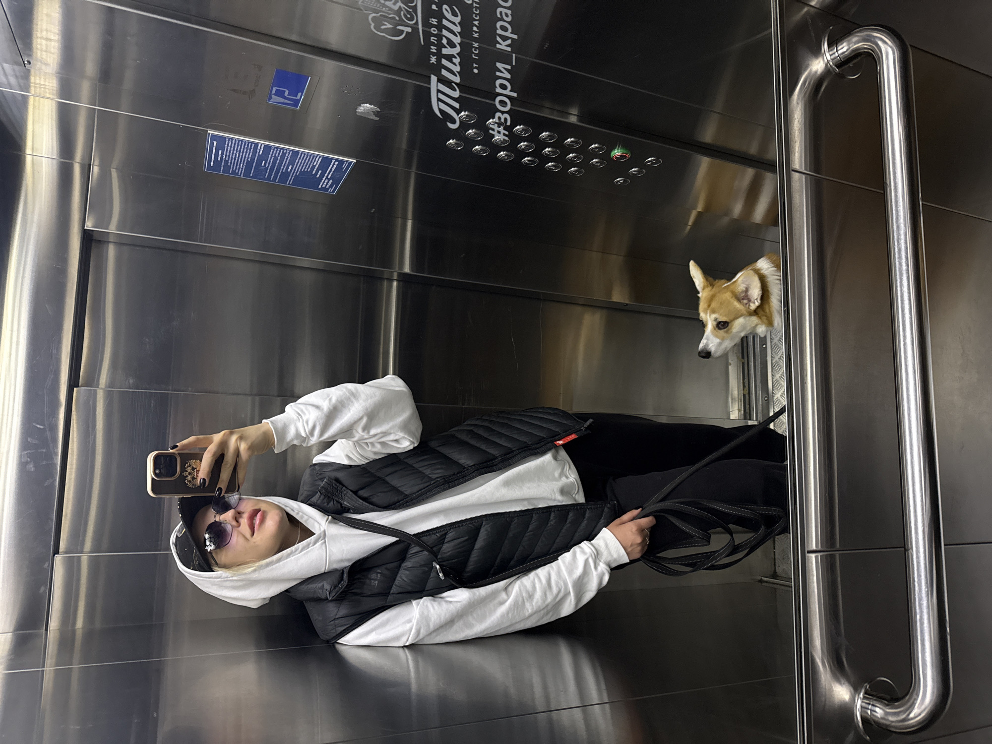 A woman is taking a selfie with her mobile phone in what appears to be an elevator. She is wearing a white hooded sweatshirt, a black puffer vest, black pants, a black baseball cap, and sunglasses. The elevator is lined with stainless steel and has a control panel with buttons and some text written in Russian. A corgi dog is visible looking through the partially open elevator door. The text "ЖИЛОЙ РАЙОН Михие #зори_крас" is visible, suggesting the location is in the city of Krasnoyarsk, Russia.