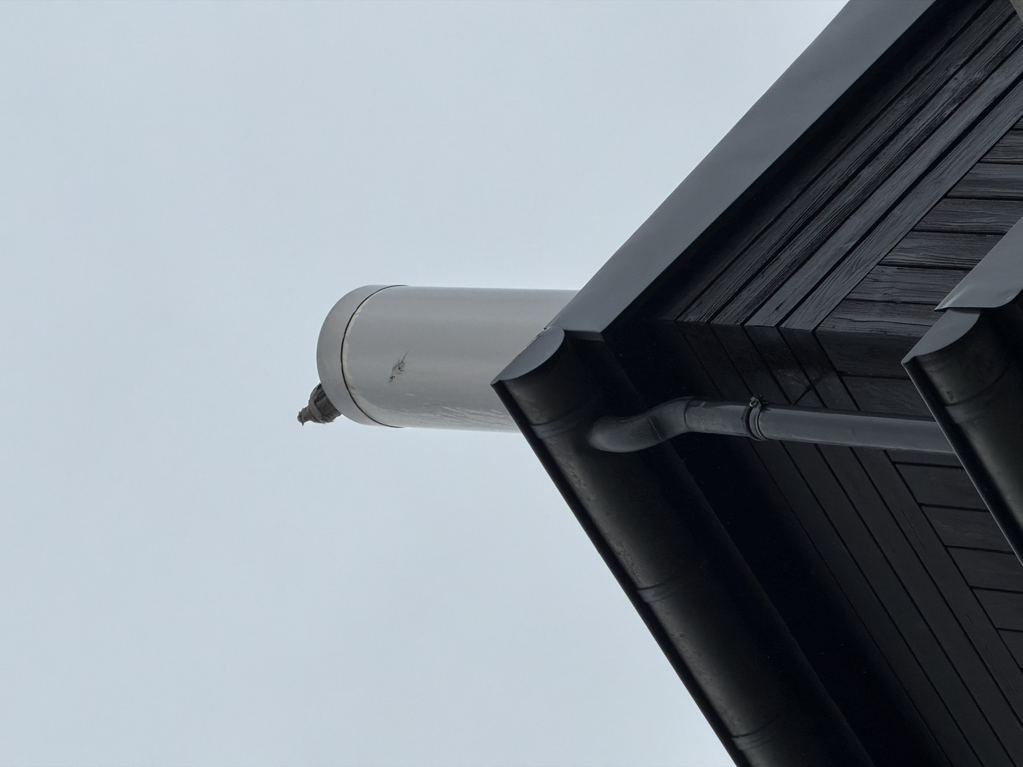 The image shows a close-up of a building's eaves, angled upwards towards a grey sky. The eaves are clad in dark, possibly black, wooden planks, arranged horizontally. A grey, cylindrical pipe extends horizontally from the building's side, seemingly venting something. A section of black guttering runs along the edge of the eaves. No people are visible. The location, Esch-sur-Alzette, Luxembourg, is not discernible from the visual content.