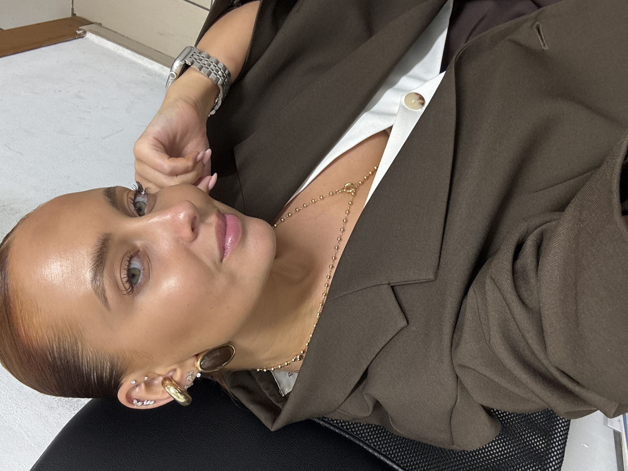 A woman with light skin, light green eyes, and auburn hair is seen from a low angle wearing a brown blazer, a white shirt, and a gold necklace and earrings. Her right hand is near her face. She is sitting in a black chair. The location is not discernible.