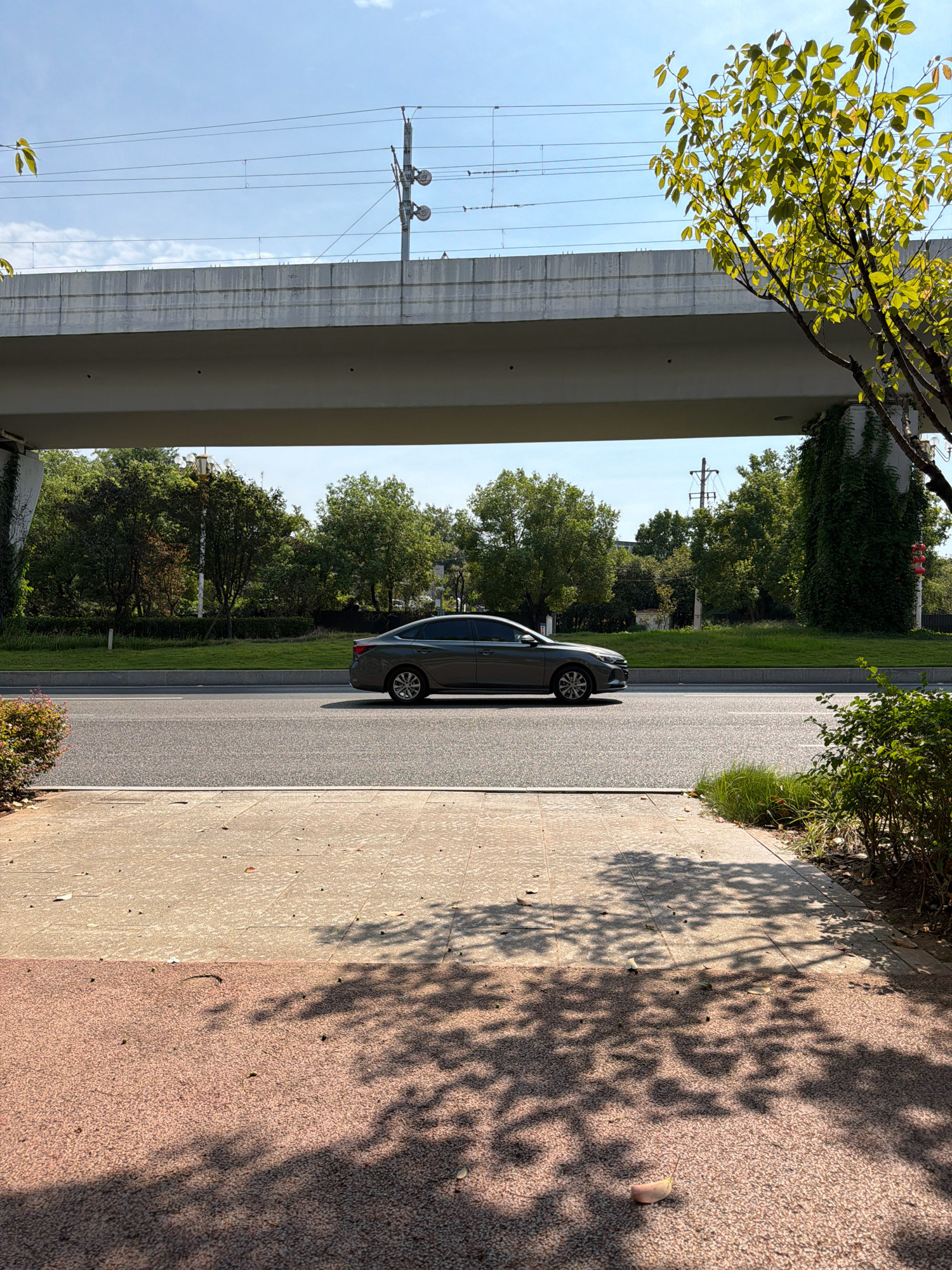 A side view captures a grey sedan driving on a paved road under an overpass. The overpass is a long, concrete structure, with train tracks visible above it. The area around the road is lined with green grass and trees, with a sidewalk in the foreground. The sidewalk has a speckled pattern with shadows of leaves cast on it. The sky is clear and blue. The scene is set in Jiangjin, China.
