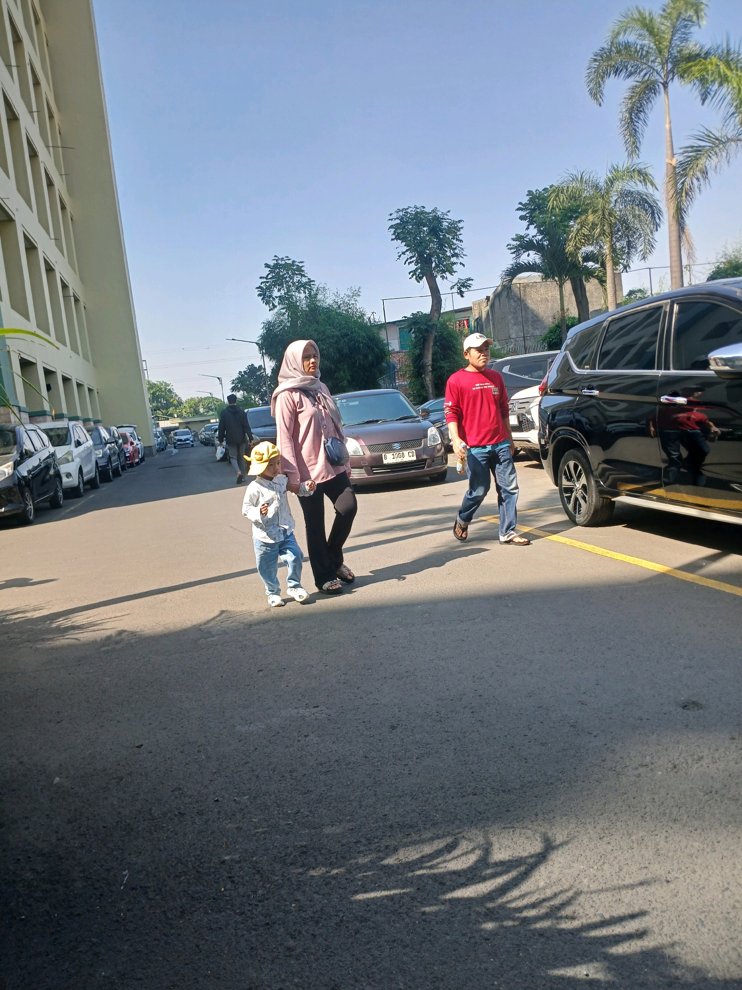 In the city of Bandung, Indonesia, on a sunny day, a family walks across a paved road with parked cars on the left and a building on the far left. A woman wearing a pink hijab and a long-sleeved shirt walks holding the hand of a young child. The child wears a yellow hat, a white long-sleeved shirt, and blue jeans. A man in a red long-sleeved shirt and blue jeans walks ahead of them, carrying a white bottle. Shadows of trees and leaves are cast on the road. A black SUV is parked on the right, and other cars are parked on the left side of the road. The background shows trees, buildings, and a clear blue sky.
