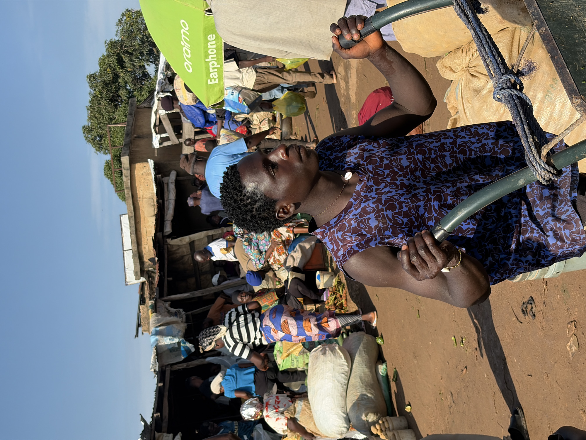 In the city of Katon Rikkos, Nigeria, a young Black man with dark skin and short, curly hair is the central subject, positioned in the foreground and facing to the right. He is wearing a sleeveless blue shirt with a brown floral pattern and a thin, light-colored necklace with a shell pendant. His right hand grasps the handle of a green wheelbarrow, while his left hand rests on its frame. A thick, blue rope is tied around the wheelbarrow's handle and a large, tan sack of produce.
Behind him, the bustling market environment unfolds. Several individuals, a mix of men and women, are engaged in various activities. Some are walking, others are standing or sitting amidst piles of goods. Bags of produce, some large and white, others smaller and tan, are stacked around the market. Makeshift stalls with corrugated metal roofs and fabric awnings line the background, suggesting a lively commercial area. A bright green awning with the text "oraimo" and "Earphone" is visible on the right side of the frame, indicating commercial advertising within the market. The ground is unpaved, appearing to be dirt. The sky above is clear and blue. The lighting suggests it is daytime, with shadows cast on the ground, indicating a sunny day.