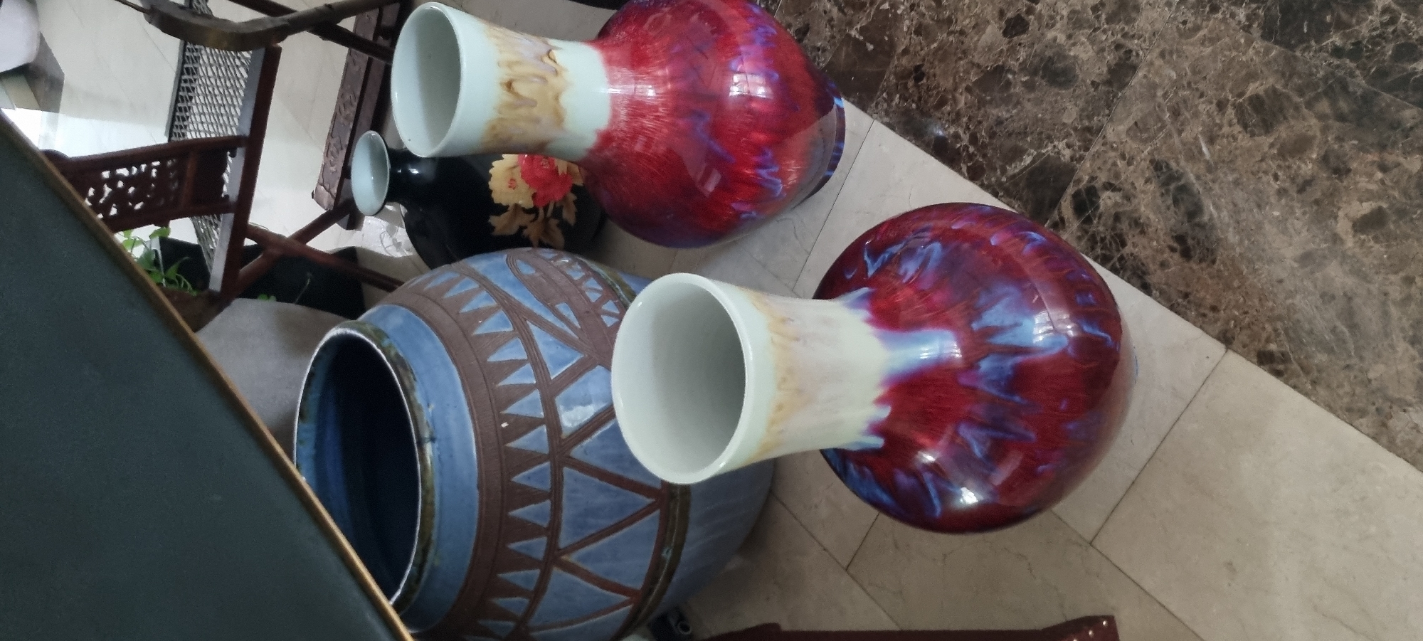 The image shows three vases, two red and one blue, arranged on a light-colored tiled floor near a dark brown faux marble wall or counter. A small black vase with floral designs sits between the large vases. The vases are cylindrical with round bodies. A portion of a dark-colored chair is visible to the left of the blue vase. The vases are near a carved wooden chair.
This is not enough data to determine the location in Falomo, Nigeria.