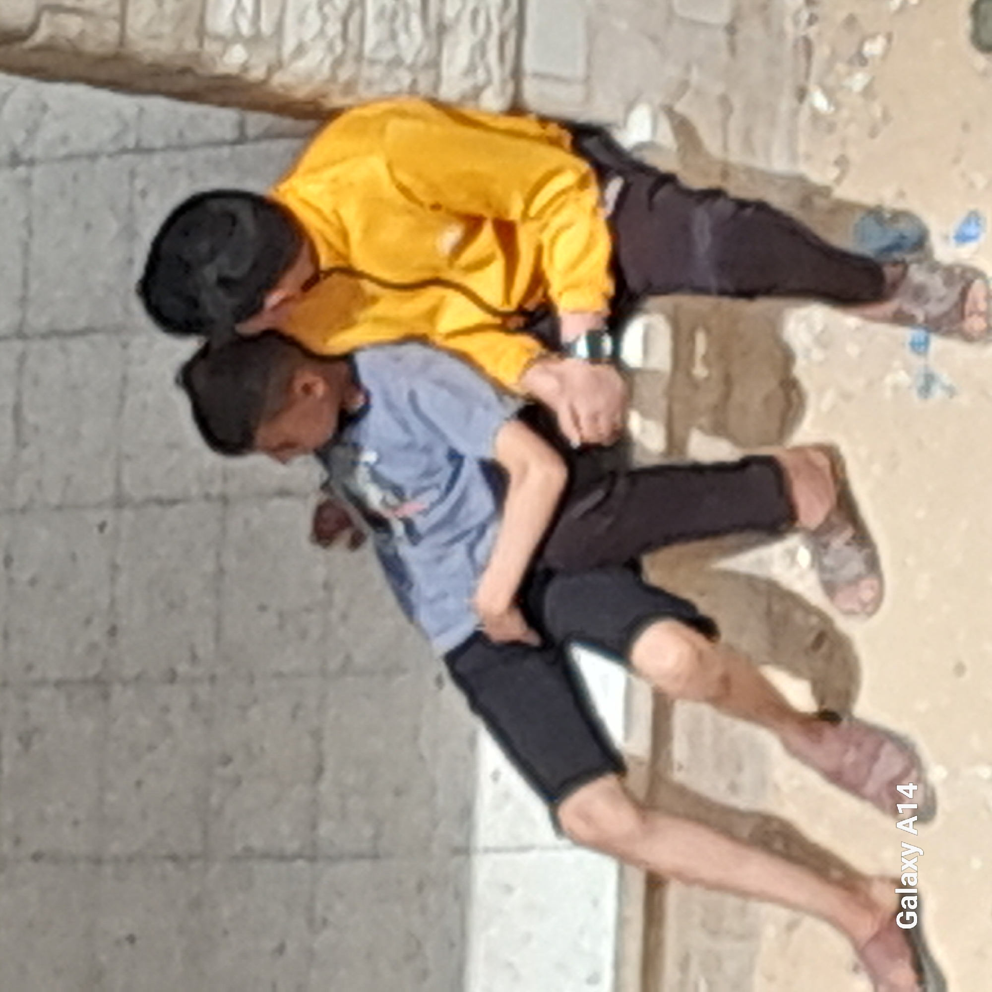 The image displays two young males in an outdoor setting, captured with a slightly soft, painterly effect. The image appears to be rotated 90 degrees clockwise; when viewed upright, the two individuals are standing or leaning.
The male on the left wears a bright yellow long-sleeved top and dark pants. He has dark hair and a wristwatch on his left wrist. He is positioned behind the second male, with his right arm extended, appearing to rest his hand on the back or shoulder of the other individual.
The second male, positioned slightly in front and to the right, wears a light blue short-sleeved t-shirt and dark shorts. He also has dark hair and is leaning forward, looking downwards. His feet are visible, wearing light-colored sandals.
The activity between the two is ambiguous; it could be a supportive gesture, a playful interaction, or simply their posture.
The background consists of a textured, light-colored wall or vertical surface on the left side, and a lighter, sandy or dirt-like ground surface on the right, suggesting an outdoor environment. The lighting indicates it is daytime.
The text "Galaxy A14" is visible vertically along the bottom right edge of the image, indicating the device used to take the photograph.