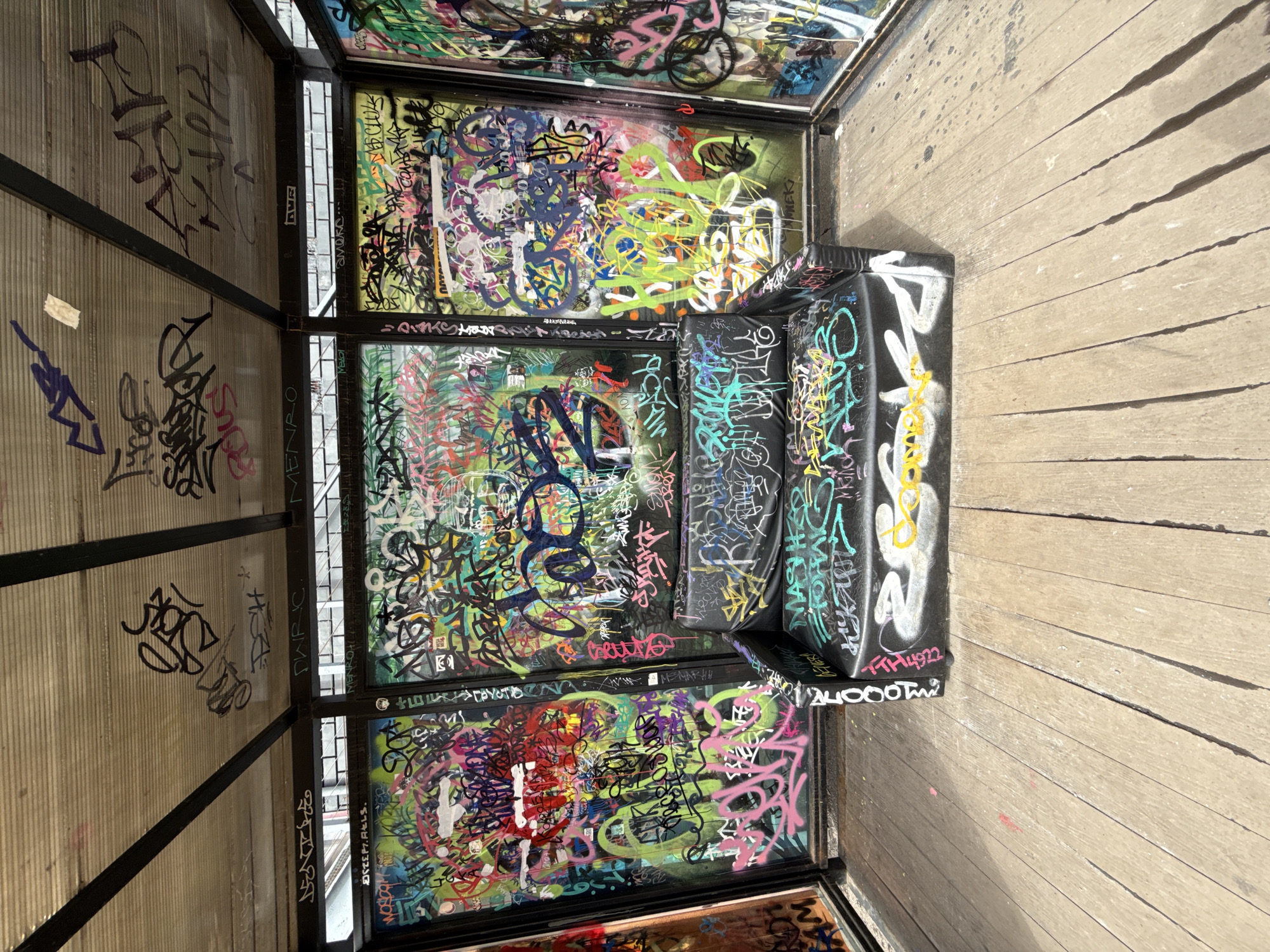 The image captures the interior of a small, heavily graffitied structure, possibly a shelter or an art installation, located in Moscow, Russia. The scene is dominated by vibrant street art covering almost every surface.
The structure features multiple framed panels; those on the left are translucent and ribbed, while others are opaque and form an enclosed space. A black, upholstered bench-like object, entirely covered in graffiti, is positioned within this space.
The graffiti is multi-layered and visually dense, showcasing a chaotic mix of tags, stylized letters, and abstract forms in a wide palette of colors including black, white, yellow, blue, green, pink, and red. The transparent panels allow for glimpses of underlying artwork, adding to the visual complexity.
On the right side of the frame, a wooden floor with distinct planks is visible, appearing worn with some minor blemishes. The lighting suggests it is daytime, providing clear illumination for the colorful details. No specific activity is taking place, presenting a static snapshot of this urban art environment.
Visible text fragments and tags include "MASSH", "DW RIC", "MENRO", "MEKI", "NOV", "STEP7.AELS.", "амека...", and "1000", interspersed among numerous other illegible markings.