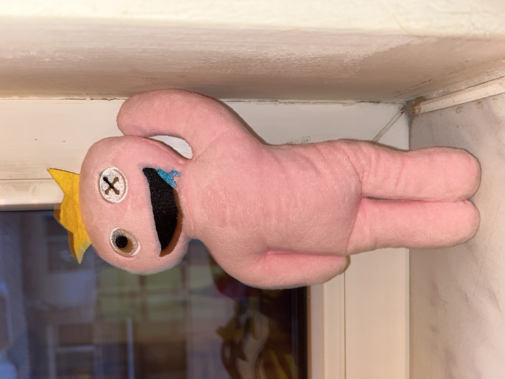 This image captures an indoor scene in Varna, Bulgaria, featuring a light pink, anthropomorphic plush toy standing upright on a light-colored surface, likely a windowsill or counter. The toy has a soft, fuzzy texture. Its head features a dark brown, circular left eye and a white right eye marked with a black "X," both outlined in white and appearing slightly reflective. A wide, open, irregularly shaped black mouth is present, with a jagged, light blue liquid-like embellishment dripping from its right side. A yellow, three-pointed crown-like accessory is positioned on top of the toy's head. Its right arm is slightly raised and bent at the elbow, while the left arm hangs down its side. The toy stands on two stubby legs.
In the background to the left, a large window reveals a blurred, low-light view of an exterior urban environment, possibly buildings or structures across a street, with indistinct warm and cool light sources suggesting an evening or night setting. The window sill directly beneath the toy is white and flat. To the right of the toy, a light-colored, possibly off-white or cream-colored, textured wall runs vertically, showing some signs of wear, minor cracking, and possibly paint inconsistencies near its corner with another perpendicular wall. At the bottom right, a narrow, light-colored wooden trim or baseboard is visible against the wall, with a thin, transparent wire or tube running near the floor level. The floor surface in the foreground is light-colored and exhibits subtle reddish-brown patterns or stains. No people or specific actions are depicted; the scene is static.