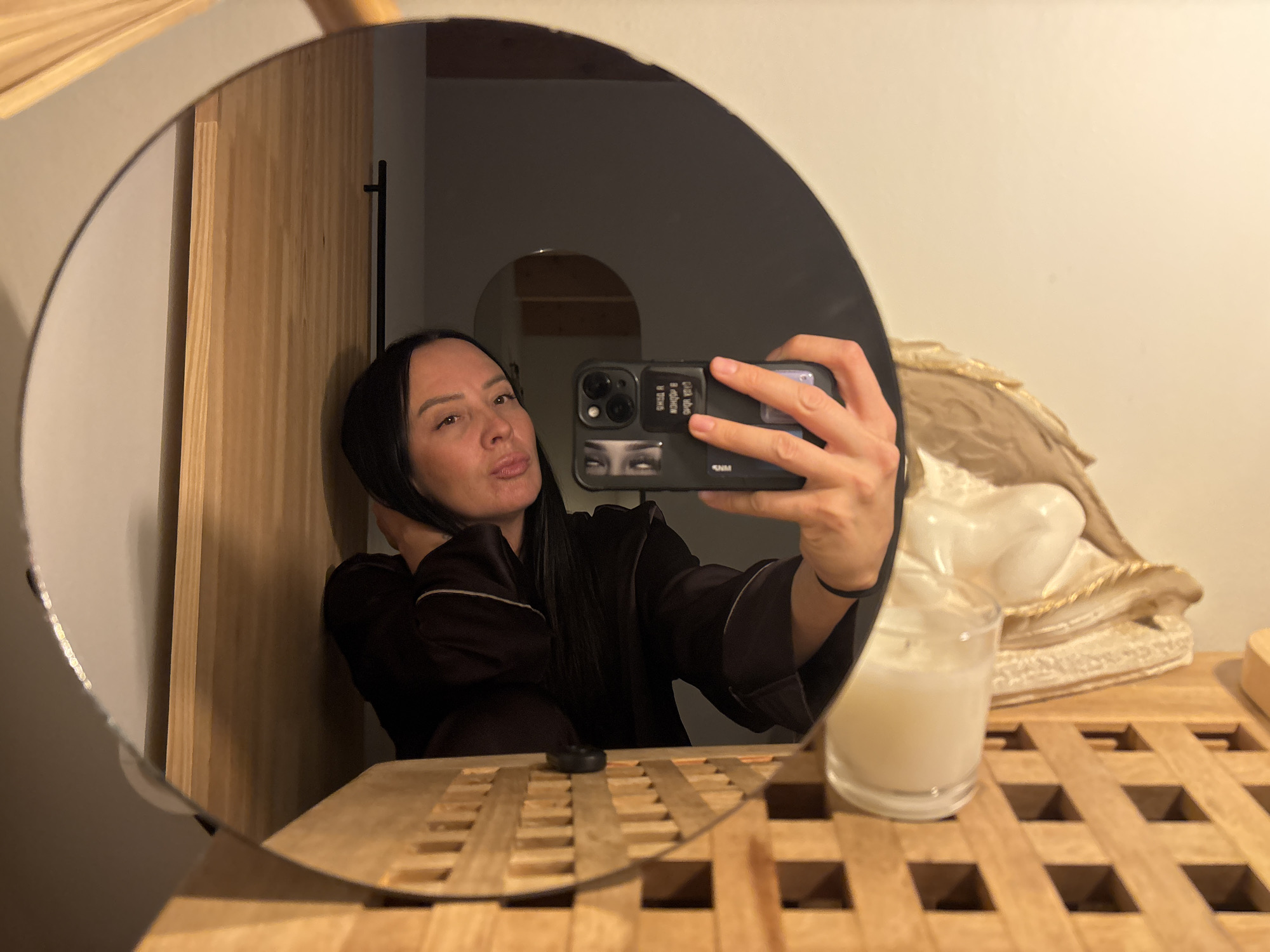 A woman is taking a selfie in a round mirror. She is wearing a dark robe with light trim. She is holding a black smartphone with stickers on the back. Her hair is dark and pulled back. The mirror reflects a bathroom with a wooden cabinet, a candle in a glass jar, and a decorative bust. In the background, there is a glimpse of another room through an arched doorway. The location is Chaikovsky, Russia.