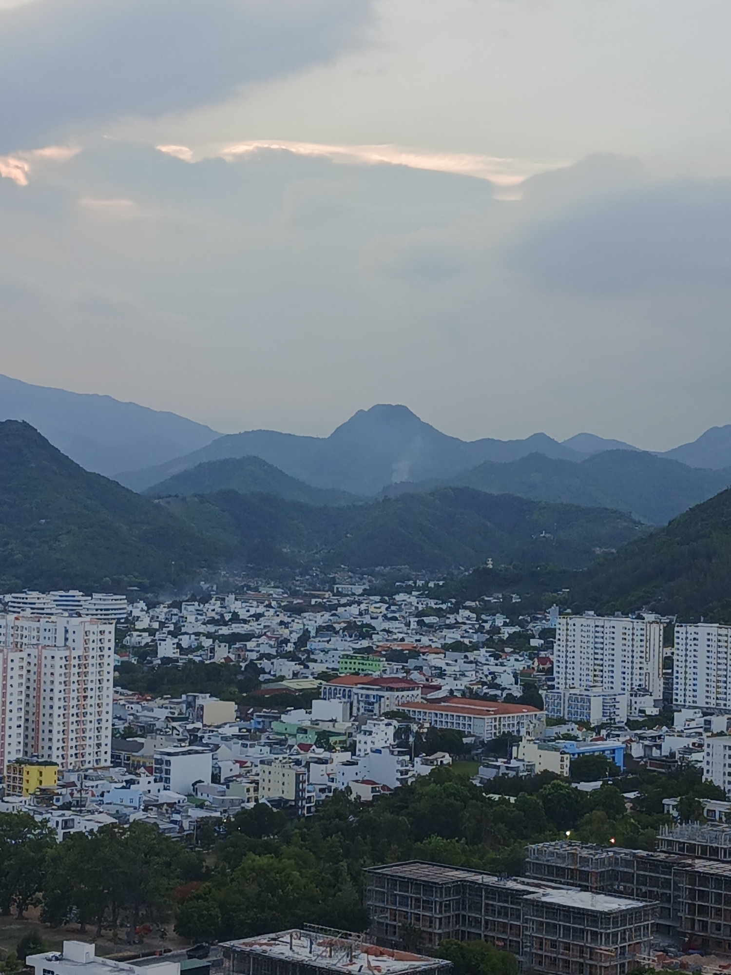This is an aerial, full shot image of a city bordering green mountains under a cloudy sky. In the foreground, there are several unfinished concrete buildings with visible rebar structures. The middle ground is a dense urban area filled with a mix of mid-rise and high-rise apartment buildings, commercial structures, and houses, characterized by their white and pastel colored facades. Lush green trees dot the cityscape, especially in the lower portions of the image, and line some of the streets. The background is dominated by layers of green, hazy mountains stretching across the horizon, with the sky above being overcast with soft, pale light filtering through the clouds. The image appears to be taken during the day, likely late afternoon or early morning, given the soft lighting. The specific location is in Bac Nha Trang Ward, Vietnam.