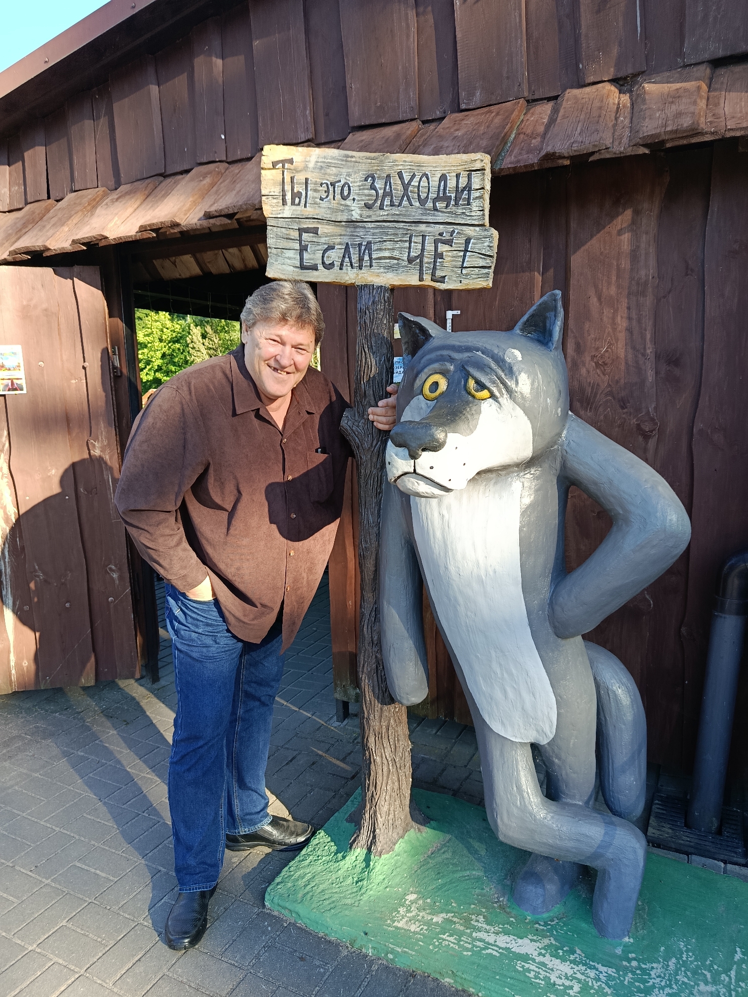 A man in a brown shirt and blue jeans stands to the left of a cartoonish grey wolf statue, leaning against a wooden post. The statue is on a green base. Behind them is a wooden structure with a sign written in Cyrillic that reads "Ты это, заходи. Если чё." (roughly translated to "Hey, come in. If anything."). The ground is paved with light-colored bricks, and sunlight casts long shadows. The background shows trees and foliage. The overall setting appears to be an outdoor recreational area or attraction, possibly in the Мяжанскі сельскі Савет region of Belarus, given the context provided.