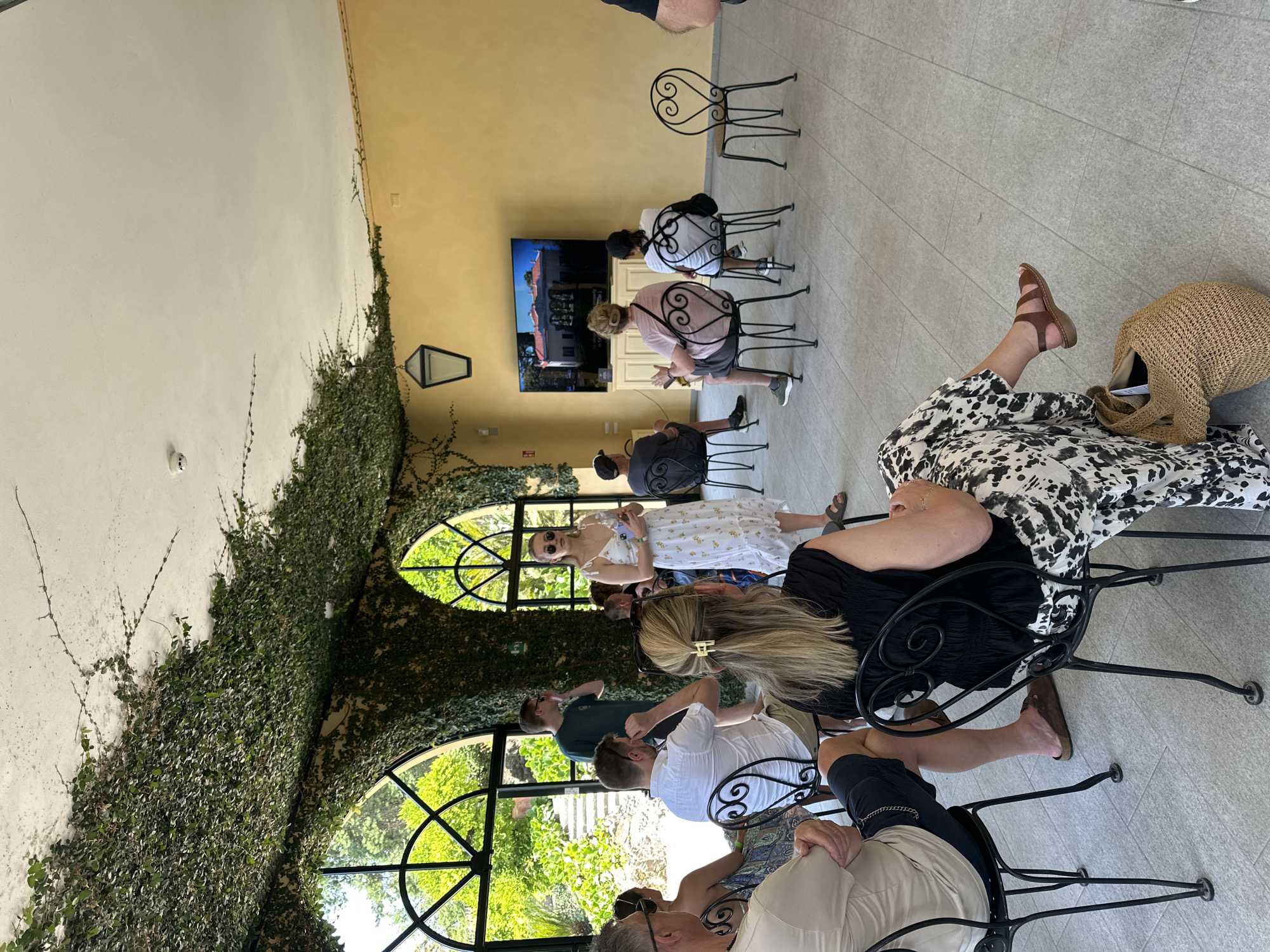In a room with a high ceiling covered in lush green ivy, a group of people are gathered. The room has large arched windows offering a view of green foliage and stairs outside. A woman in a white dress with a floral pattern stands in the center, holding a phone, addressing the seated audience. The audience members, mostly facing away from the camera, are seated in black metal chairs. A large flat-screen television on a stand displays an image of a house. The walls are painted a warm yellow, and the floor is tiled with light grey squares. The scene appears to be taking place in Tremezzina, Italy, possibly at a historical villa or a venue with a similar architectural style.