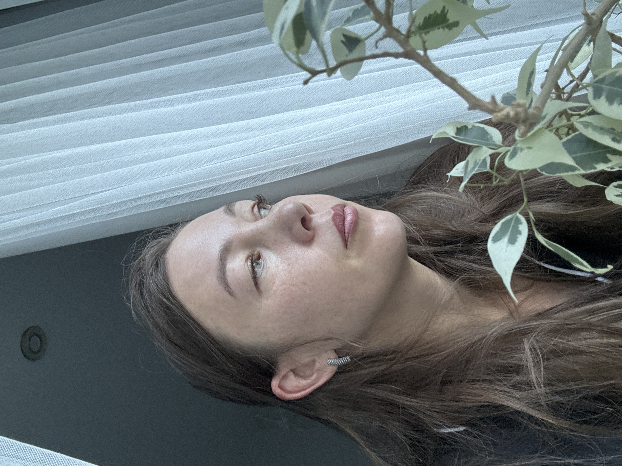 This close-up portrait, oriented 90 degrees counter-clockwise, features a young woman indoors in Chaikovsky, Russia. Her head is tilted upwards and to the right, with her gaze directed slightly off-frame, conveying a thoughtful or serene expression. She has long, dark brown hair, light brown or hazel eyes, and a scattering of faint freckles across her nose and cheeks. Her lips show a subtle pinkish-red color. A textured, silver-toned ear cuff or helix piercing adorns her right ear.
In the upper right corner, a branch of an indoor plant, possibly a variegated ficus with green and cream-colored leaves, extends into the frame. The background consists of light-colored, pleated blinds or sheer curtains, suggesting a window. Below the woman's head, a dark, plain surface, likely a wall or furniture, is visible, featuring a small, dark circular fixture embedded within it.
The lighting is soft and natural, indicating daytime conditions. There is no explicit activity or event taking place beyond the woman's contemplative pose. No text is visible in the media file.