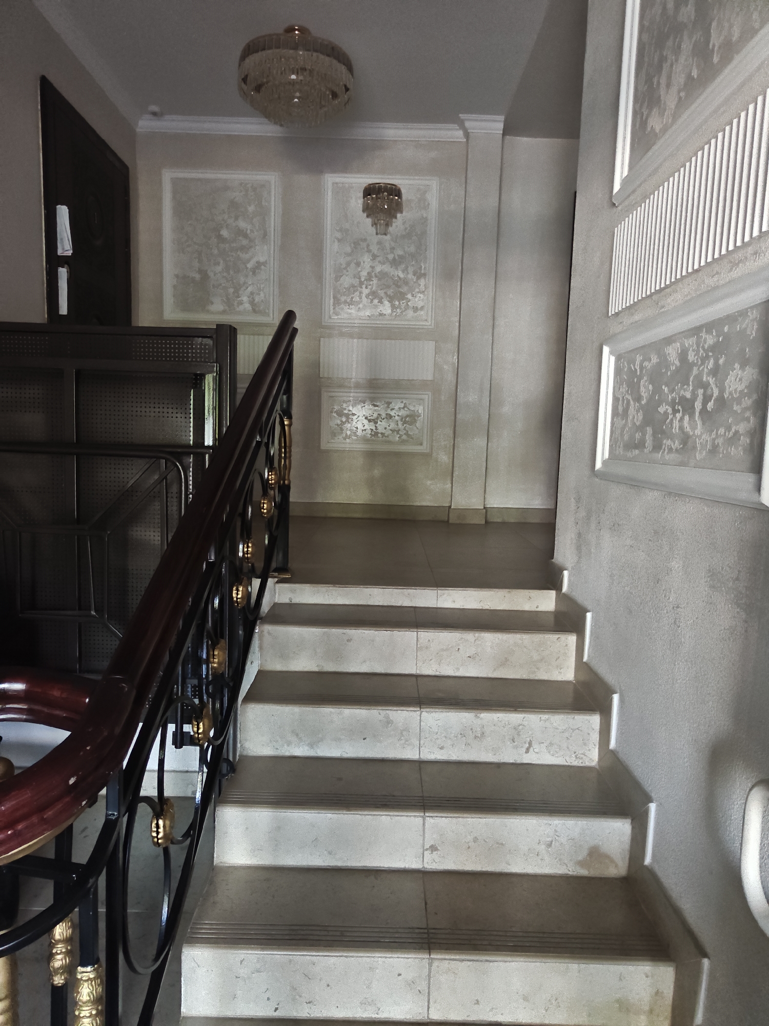 The image shows an indoor staircase with a dark wood and black metal railing, leading upwards. The stairs are made of light-colored stone or tile. The walls are decorated with textured wallpaper and white molding. Two chandeliers are visible, one at the top of the staircase and another higher up on the ceiling. To the left, there is a dark wooden door and part of a dark metal shelving unit. The environment appears to be an interior hallway or entryway, likely in a residential or commercial building in Petropavl, Kazakhstan, given the context of the prompt. No people are visible in the image. The lighting is dim, suggesting an indoor setting without direct sunlight.