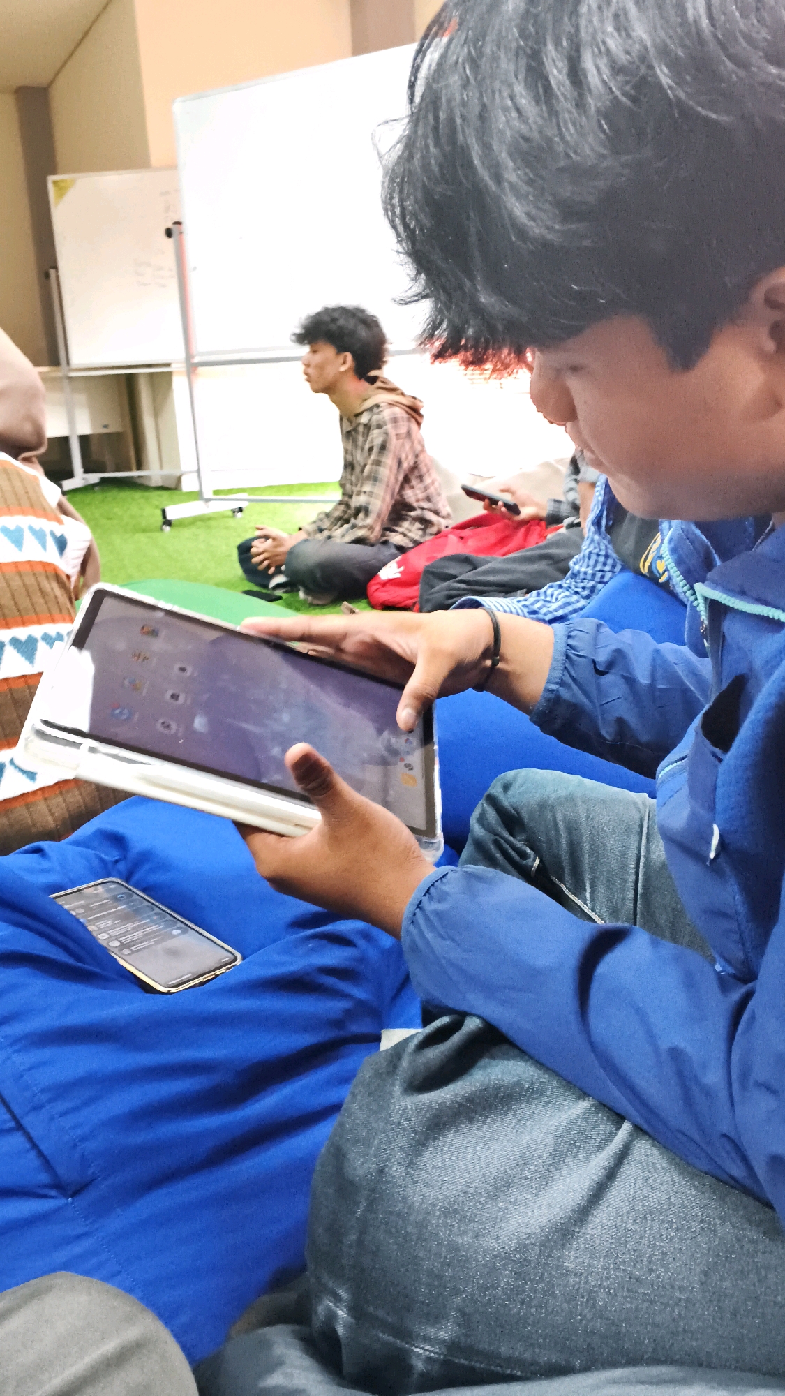 The image depicts an indoor scene, possibly a classroom or meeting room, with a green carpeted floor. In the foreground, a person is holding a tablet and a smartphone. The tablet, encased in a white cover, displays various app icons on its screen. A gold-colored smartphone lies on a blue beanbag chair. In the mid-ground, another person is seated on the floor, looking to the left. Behind them, a whiteboard is visible, suggesting an educational or presentation setting. The overall atmosphere appears casual and informal. No specific landmarks or contextual clues indicate the location within Lohbener, Indonesia, beyond the general interior setting.