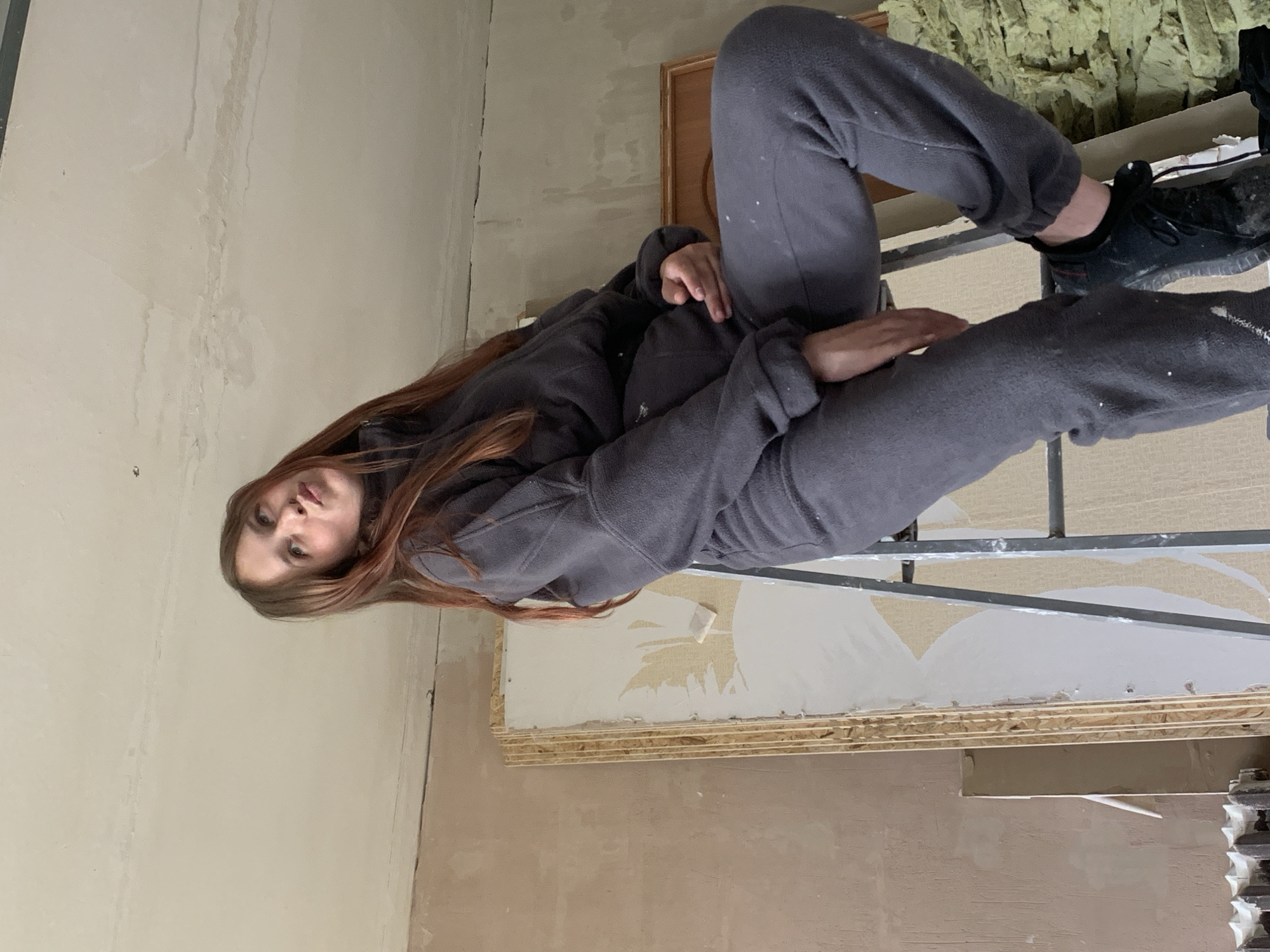 A woman in a gray sweatsuit and black sneakers sits on a metal scaffolding indoors. The walls are unfinished and show signs of repair. Insulation material is visible behind her. The location in the city of Фоки, Russia is not discernible from the image.