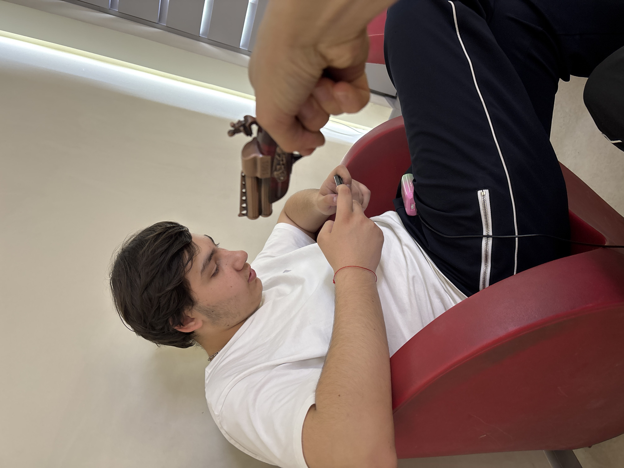 A medium-angle shot captures a person lying back in a red chair, engrossed in a small electronic device held in their hands. The individual is wearing a white t-shirt and black track pants with a white stripe along the side. They have dark hair and appear to be either resting or in a reclined position.
An older male hand with a closed fist holding a dark wooden carved doll is positioned above the person in the chair, seemingly about to drop the doll onto the device. The scene is indoors, with a light-colored wall and blinds visible in the background. A glimpse of what appears to be a window is also present.
The location is not identifiable within the image itself; therefore, I cannot confirm it to be Yerevan, Armenia.