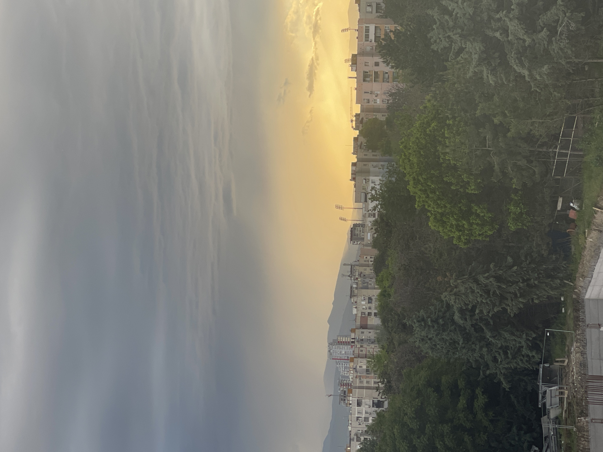 The image presents a vertically oriented cityscape, showing a contrast between weather conditions on either side of the frame.
To the left, the sky is dominated by gray, overcast conditions, possibly indicating rain or a storm. To the right, the sky transitions to a golden hue, suggestive of either sunrise or sunset.
Below the sky, a series of buildings are visible, presumably residential or commercial. Their architecture appears fairly uniform, typical of urban structures. Behind the buildings, a mountain range is faintly visible, further defining the landscape.
The foreground on the right side of the image consists of dense greenery, possibly trees or bushes. There's also some construction-related materials and equipment visible at the very bottom-right of the frame, hinting at building activity in the area.
The presence of buildings and the surrounding natural environment suggest a suburban or urban setting.
Given the prompt, it's reasonable to assume this photograph was taken in Plovdiv, Bulgaria, although specific landmarks are not distinctly visible within the image to confirm this definitively.