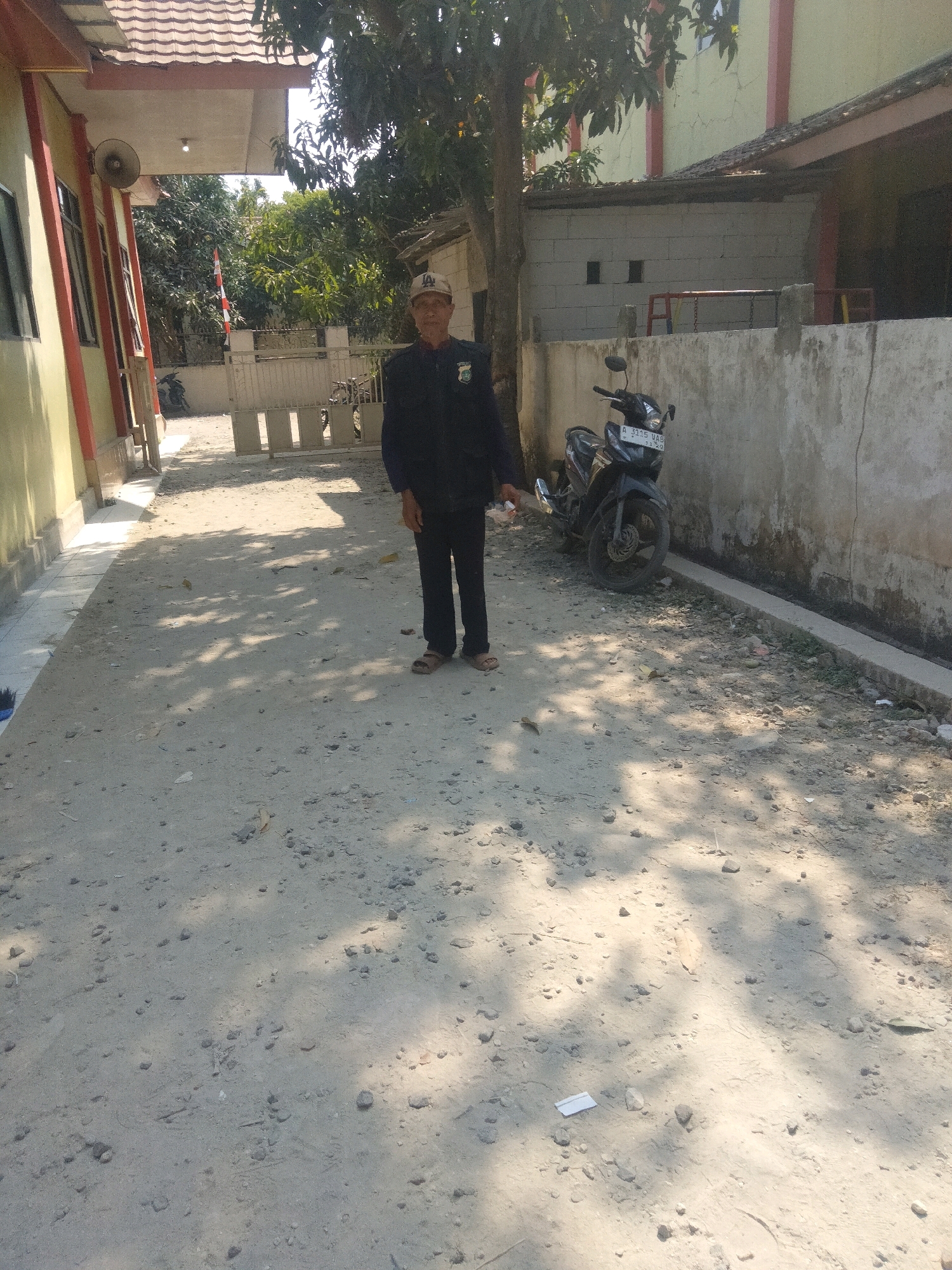 A man stands in a dirt courtyard in front of a building. The man is wearing a dark jacket and pants, a light-colored baseball cap, and sandals. A dark motorcycle is parked to the right of the man. The building appears to be a house with a tiled roof and a light-colored exterior. The courtyard is covered in dirt and small stones, with shadows cast by trees. There is a fence in the background. The scene appears to be located in Suka Sari, Indonesia.