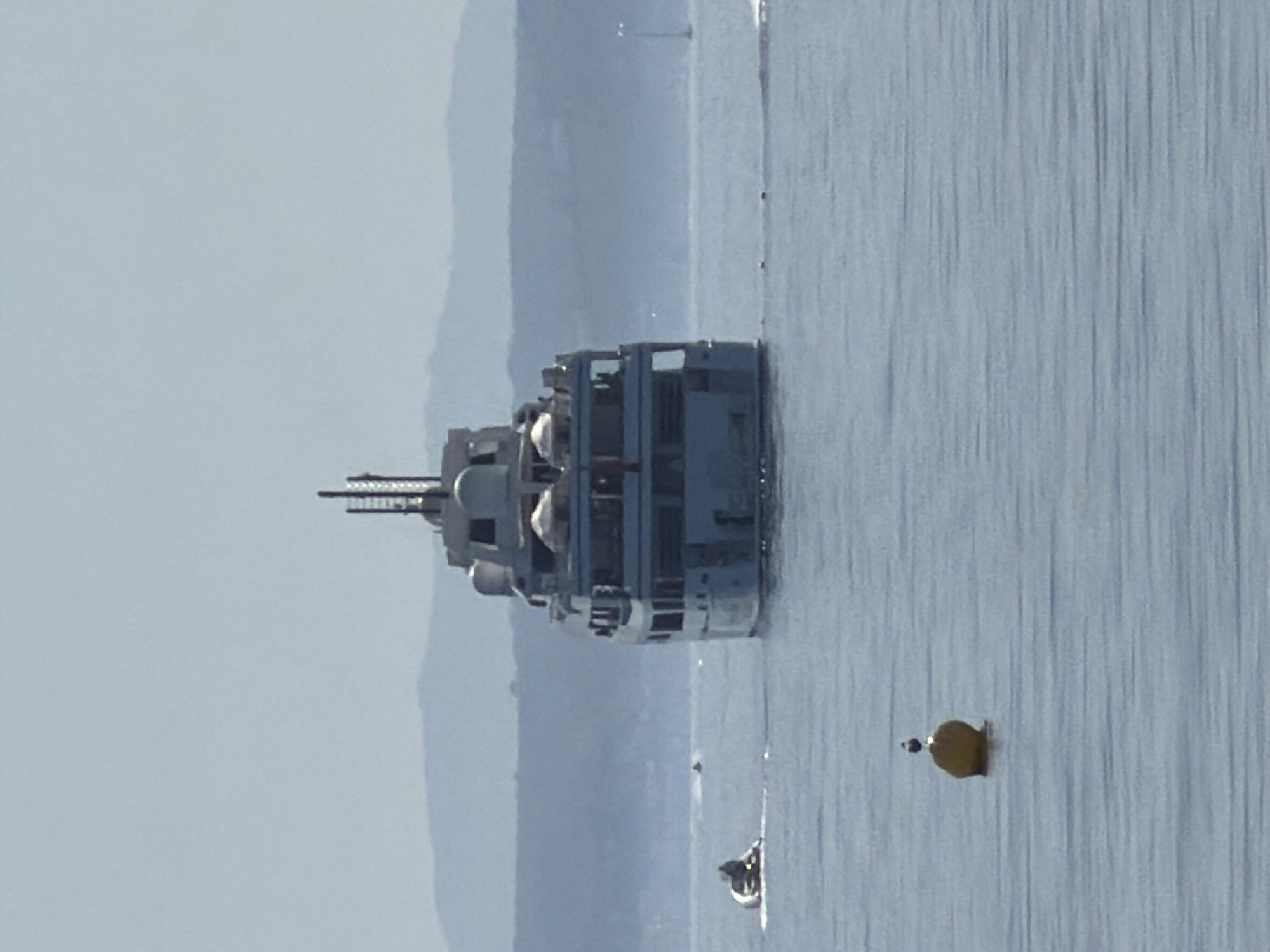 In the waters off the coast of Estepona, Spain, a large, white superyacht is anchored. The yacht, which is seen from the stern, has multiple decks and a distinctive antenna structure at its top. In the foreground, a yellow buoy with a bird perched on top floats on the calm blue water. To the left of the yacht, a small inflatable boat with two people in it is moving across the water. In the background, a hazy, distant coastline with rolling hills is visible under a clear, pale blue sky.