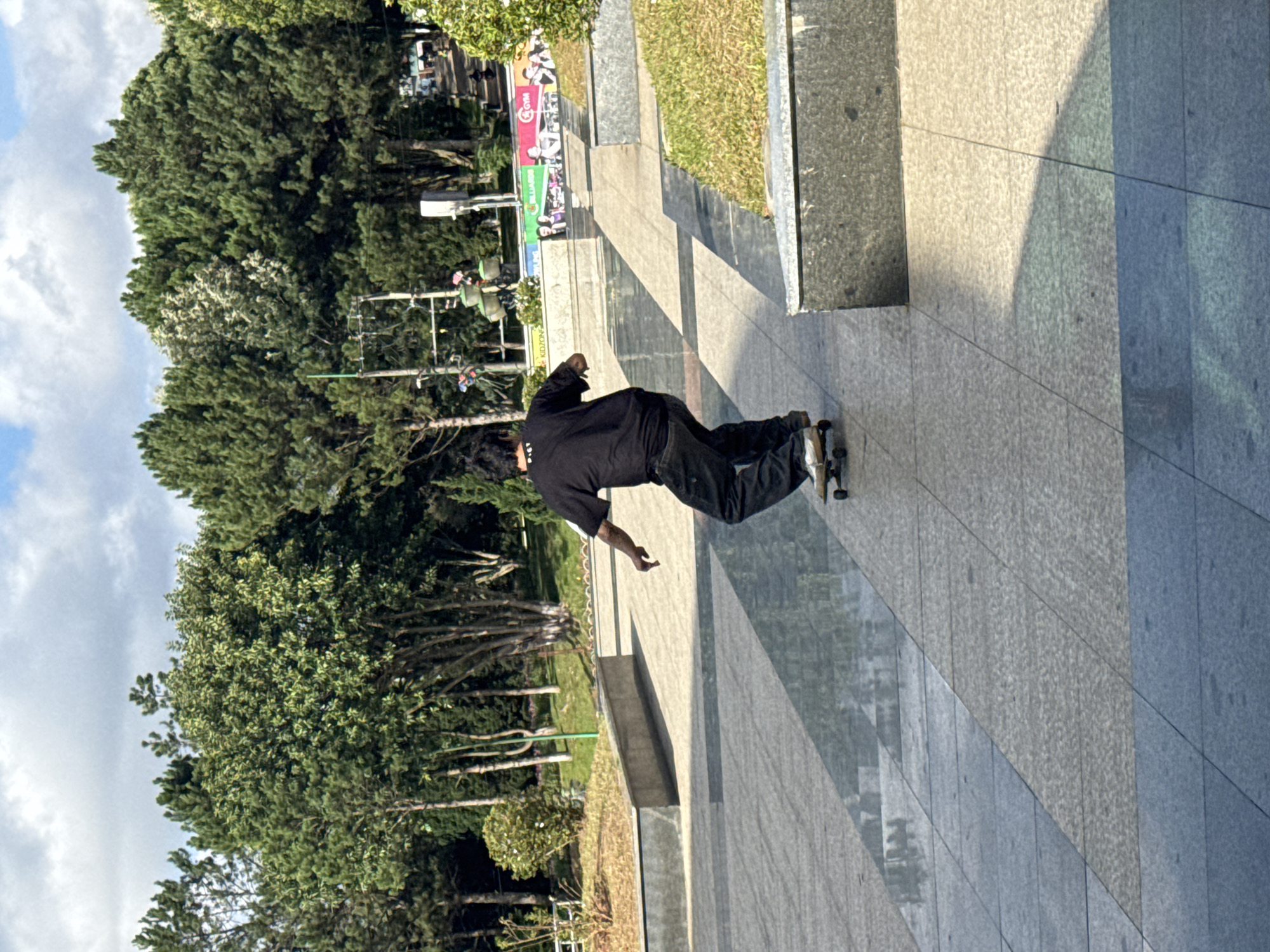 A young man is skateboarding on a paved area with a reflective surface. He is wearing a black t-shirt and dark pants. The skateboard is visible beneath his feet. In the background, there are trees and a cloudy sky. The location appears to be a park or public space in Da Lat, Vietnam, characterized by its lush greenery and architectural elements like concrete ledges.