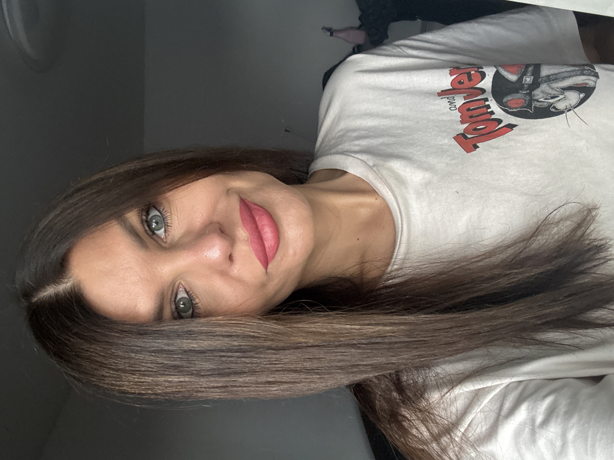 A woman with long, dark brown hair and green eyes, wearing a white t-shirt with a "Tom and Jerry" graphic, poses for a selfie. Her makeup includes pink lipstick and eyeliner. The background appears to be an indoor setting with a dark wall and a hint of a window with light coming through. The image does not provide any information about the location in the city of Chaikovsky, Russia.